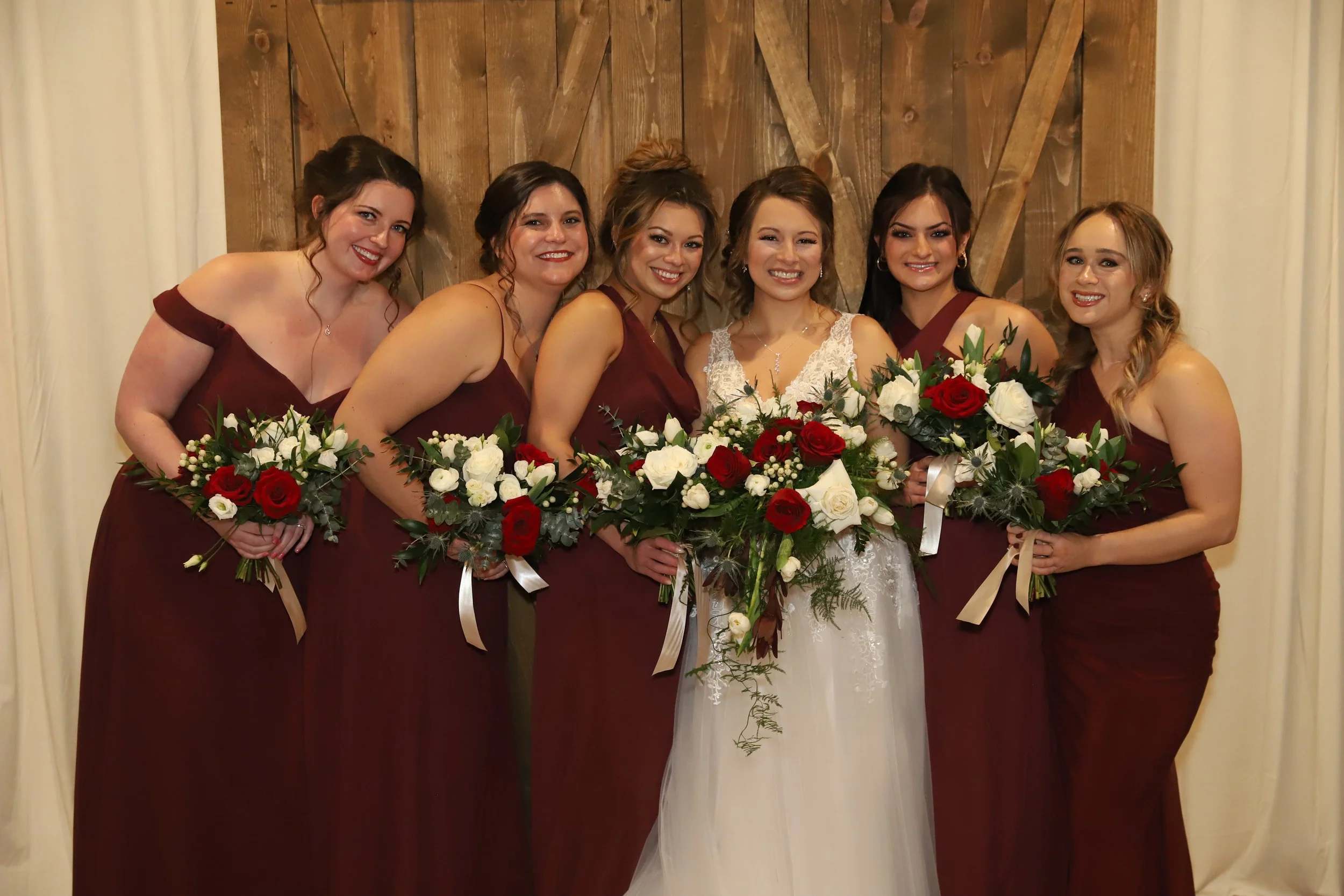 A bride and six bridesmaids standing in front of a wooden backdrop, holding bouquets of red and white flowers with greenery, smiling for the photo at a wedding.
