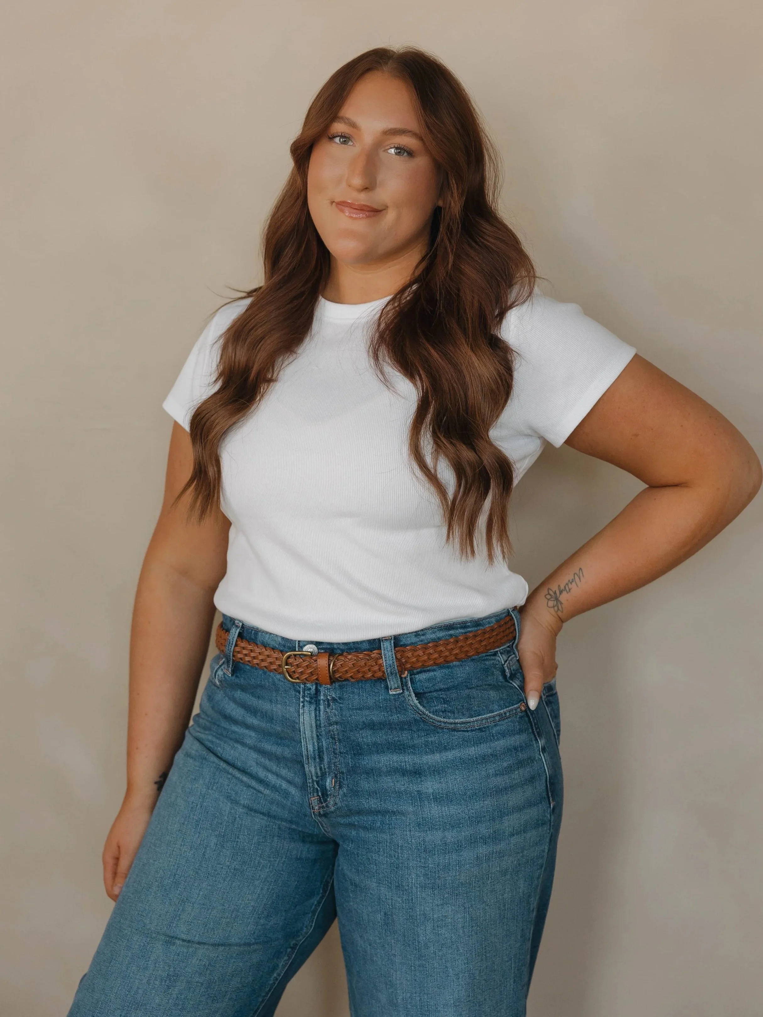 Woman with long wavy brown hair wearing a white t-shirt and blue jeans, standing against a neutral background, with one hand on her hip