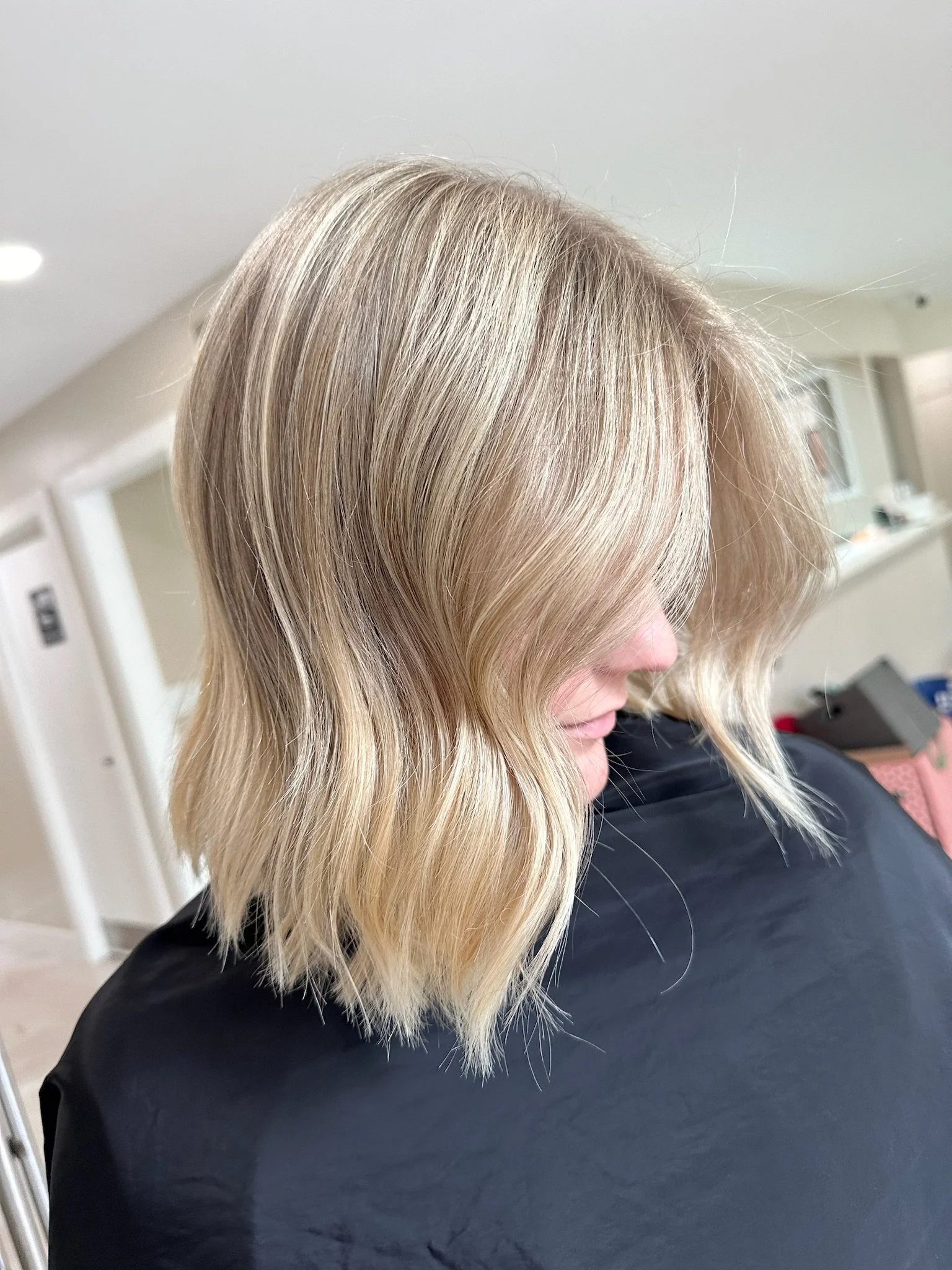 A woman with blonde, wavy hair sitting in a salon or medical setting, wearing a black cape.