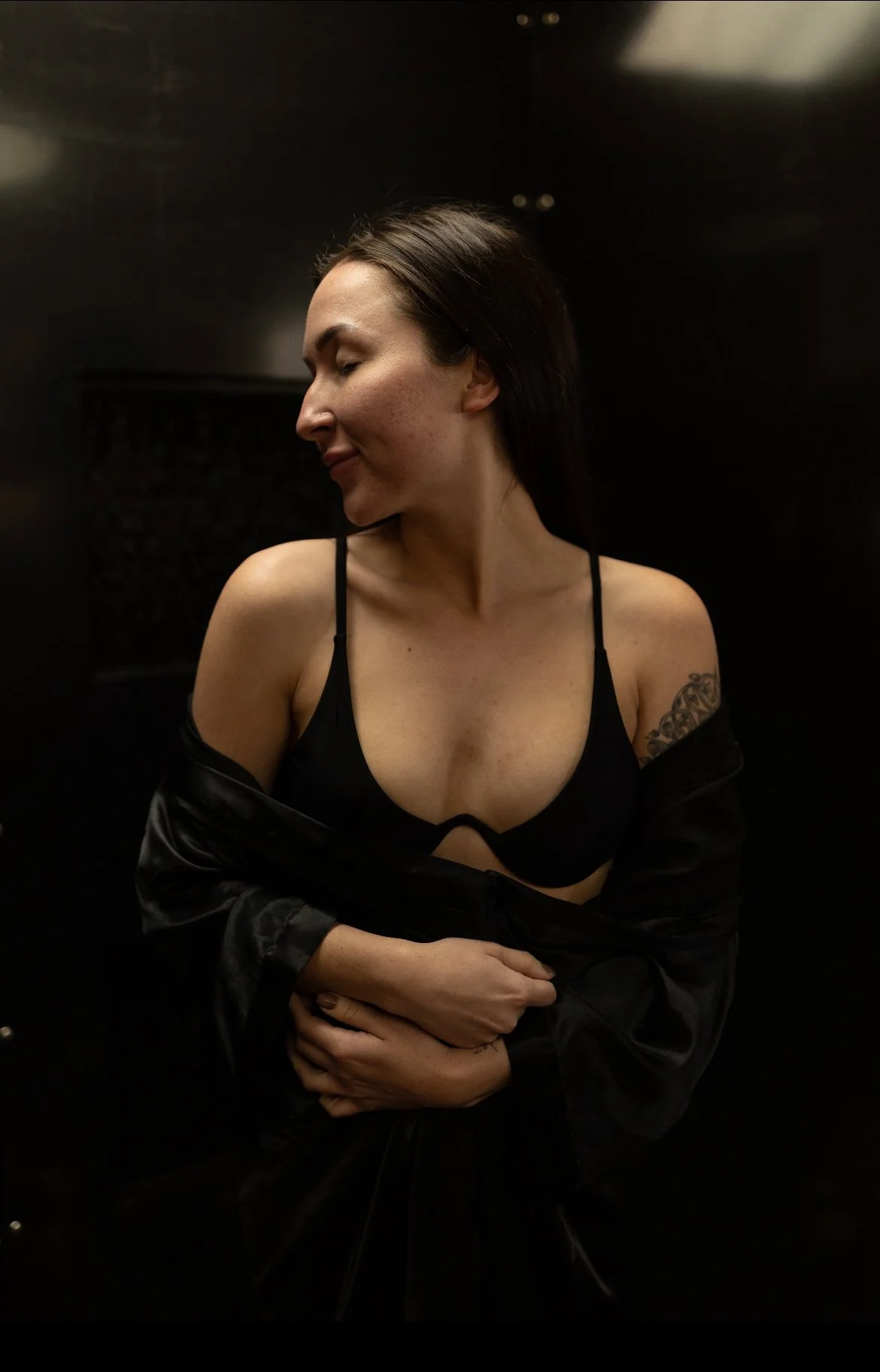 A woman with dark brown hair, wearing a black bra and a black satin robe, is standing against a dark background. She is looking down to her left and has her arms crossed over her lower abdomen.