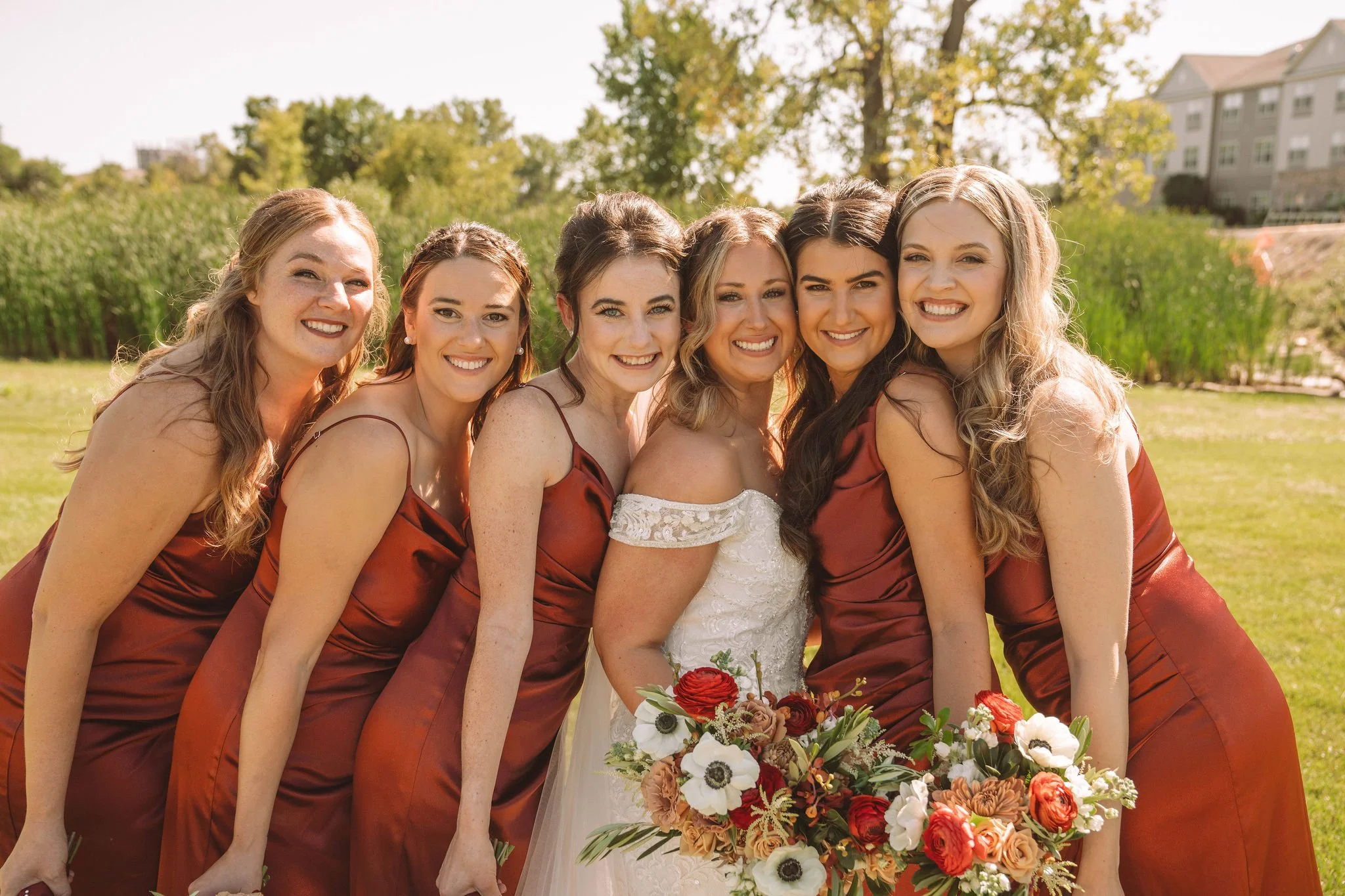 A bride in a white lace wedding dress holding a colorful bouquet stands with six bridesmaids in matching rust-colored dresses outdoors on a sunny day.