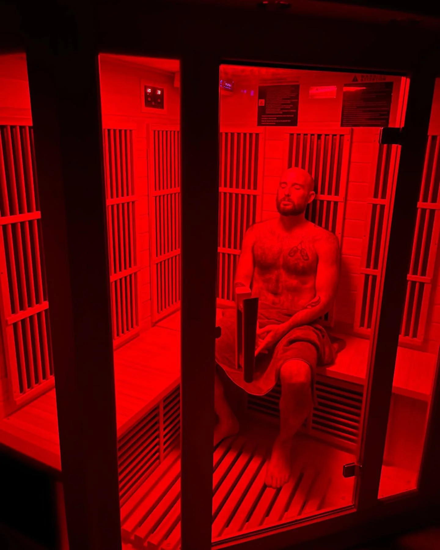 A man sitting inside a red-lit infrared sauna, surrounded by wooden panels and slats, with a towel on his lap and tattoos on his chest and arms.