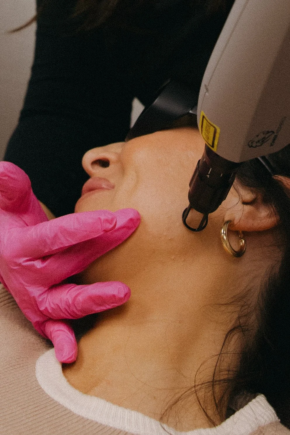 A woman receiving a cosmetic treatment near her jawline with a professional using a device, wearing pink gloves, and gold hoop earrings.
