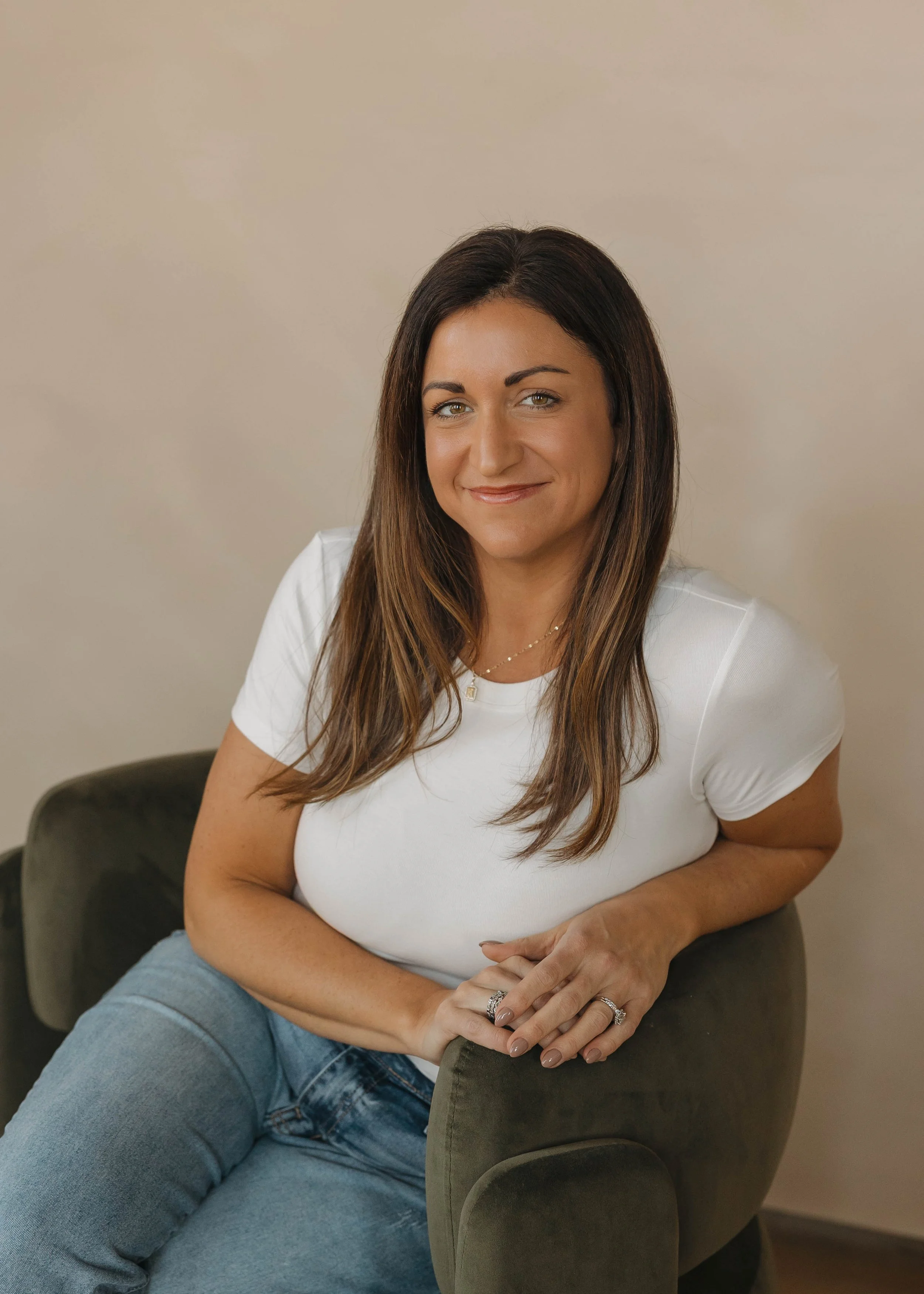 A woman with long brown hair wearing a white t-shirt and blue jeans, sitting on a dark green armchair, smiling at the camera against a beige background.