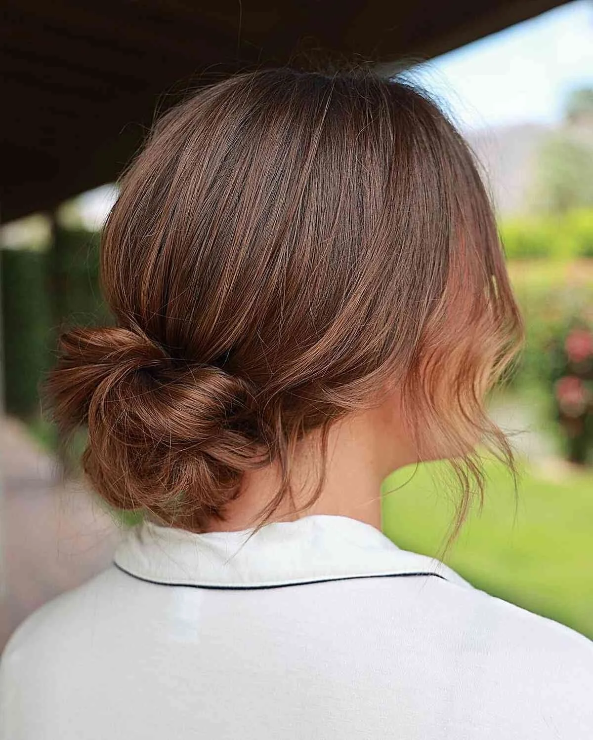 Back view of a woman with brown hair styled in a low bun and some loose waves, wearing a white shirt with a black trim, outdoors with greenery in the background.