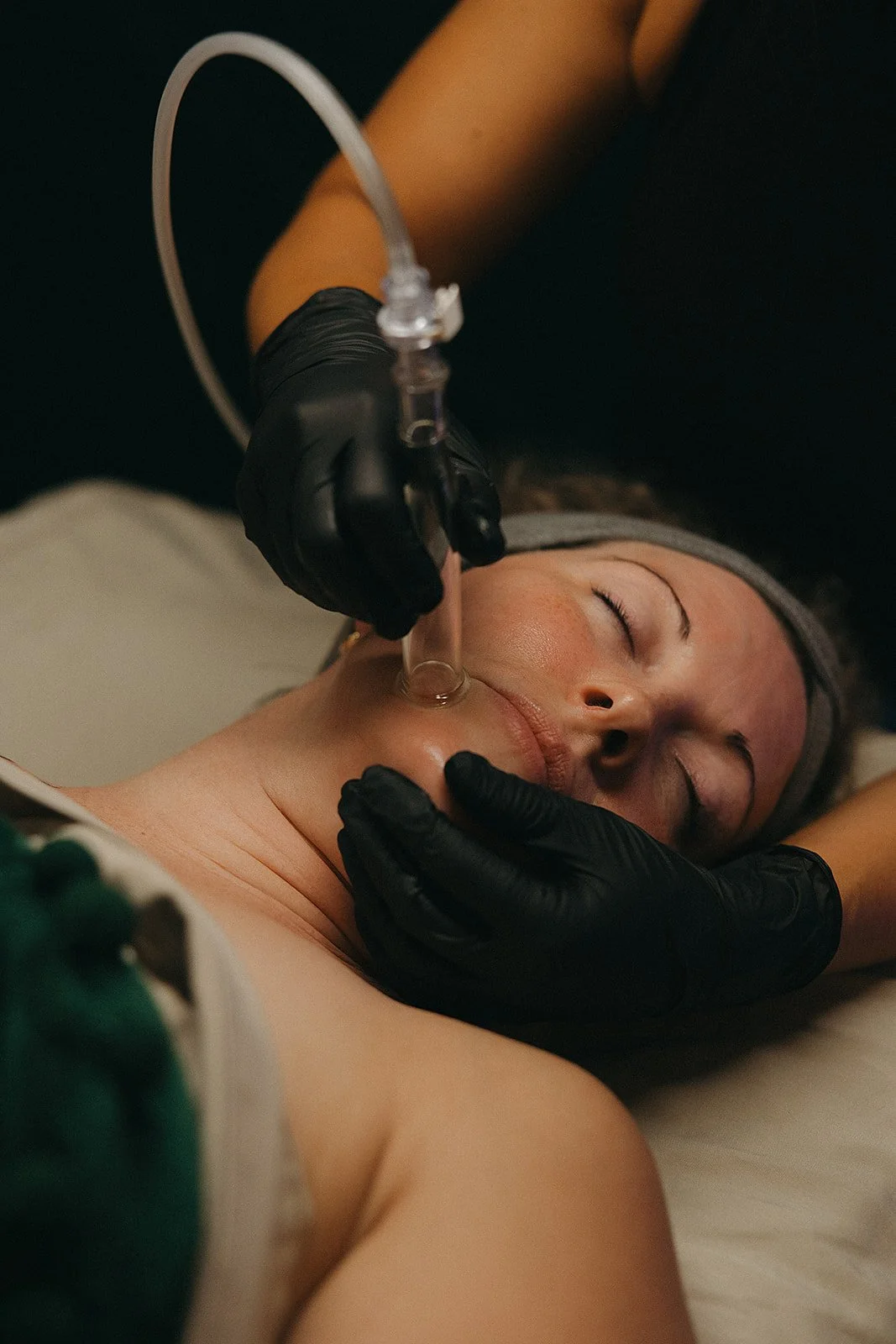 A woman lying face up with her eyes closed receives a facial treatment with a glass tool from a practitioner wearing black gloves.