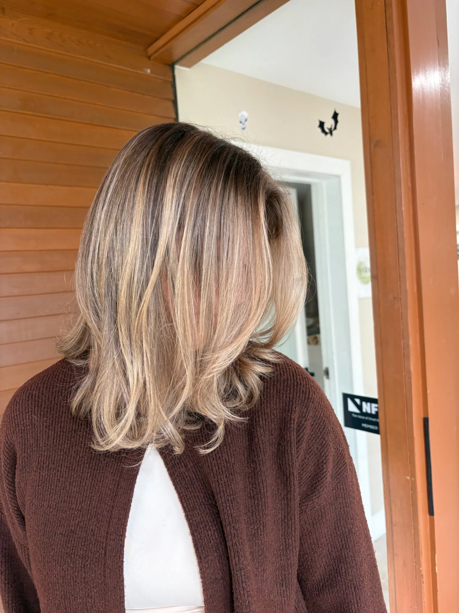 A woman with shoulder-length blonde hair, wearing a brown sweater over a white top, standing near a wooden door with Halloween decorations on the wall behind her.