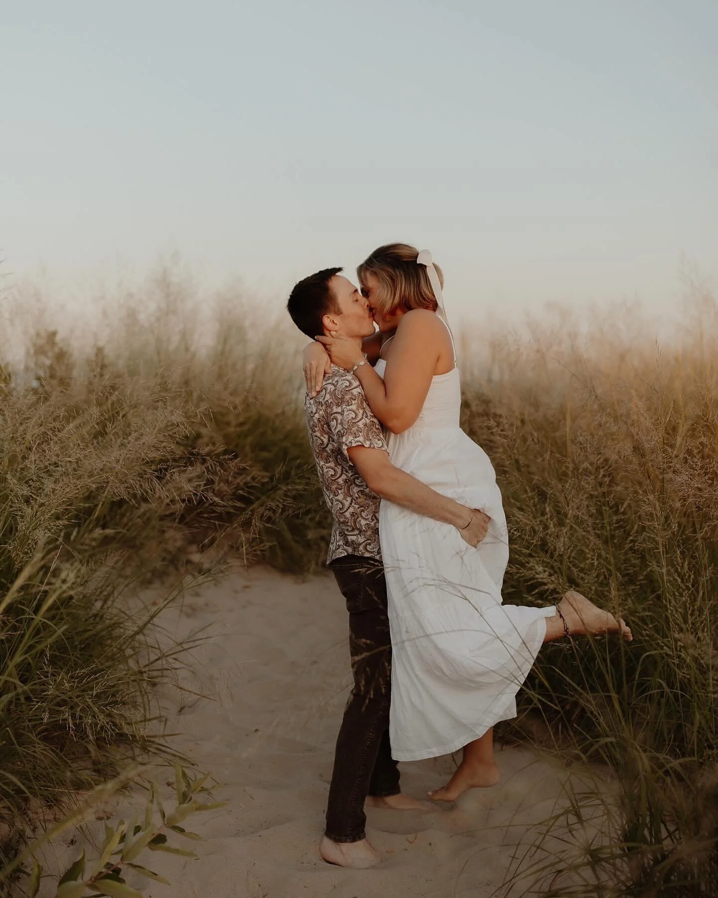 A couple sharing a kiss on a sandy path surrounded by tall beach grass during sunset.