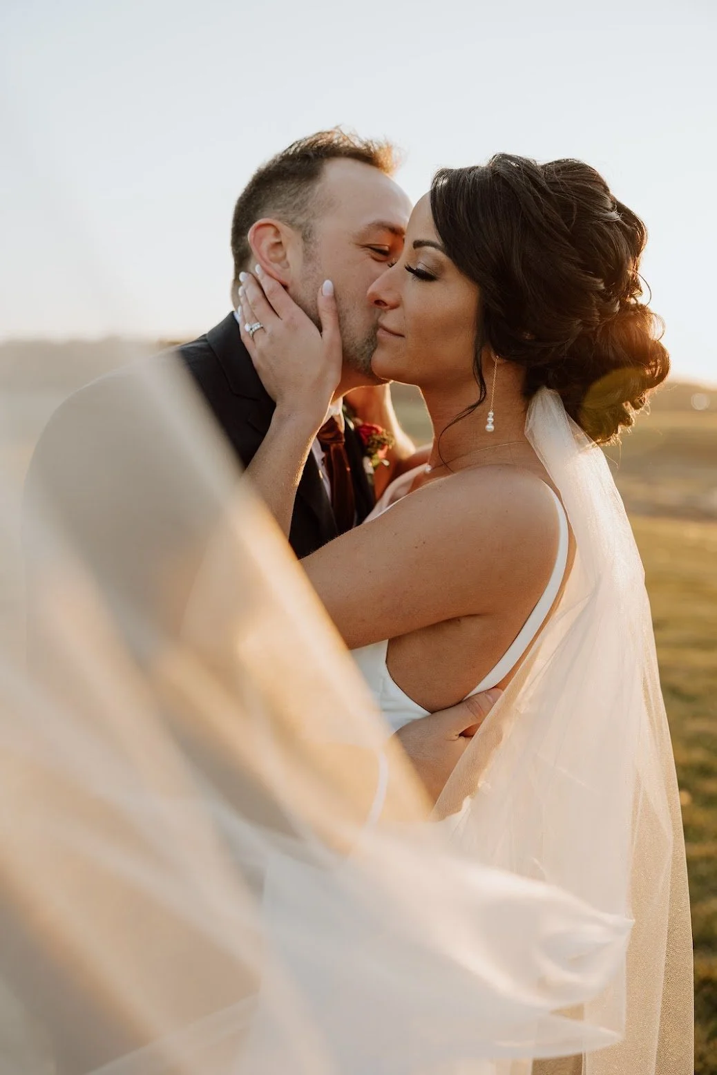 A bride and groom sharing a kiss outdoors during sunset, with the groom holding the bride's face and the bride gently holding his arm, both dressed in wedding attire.