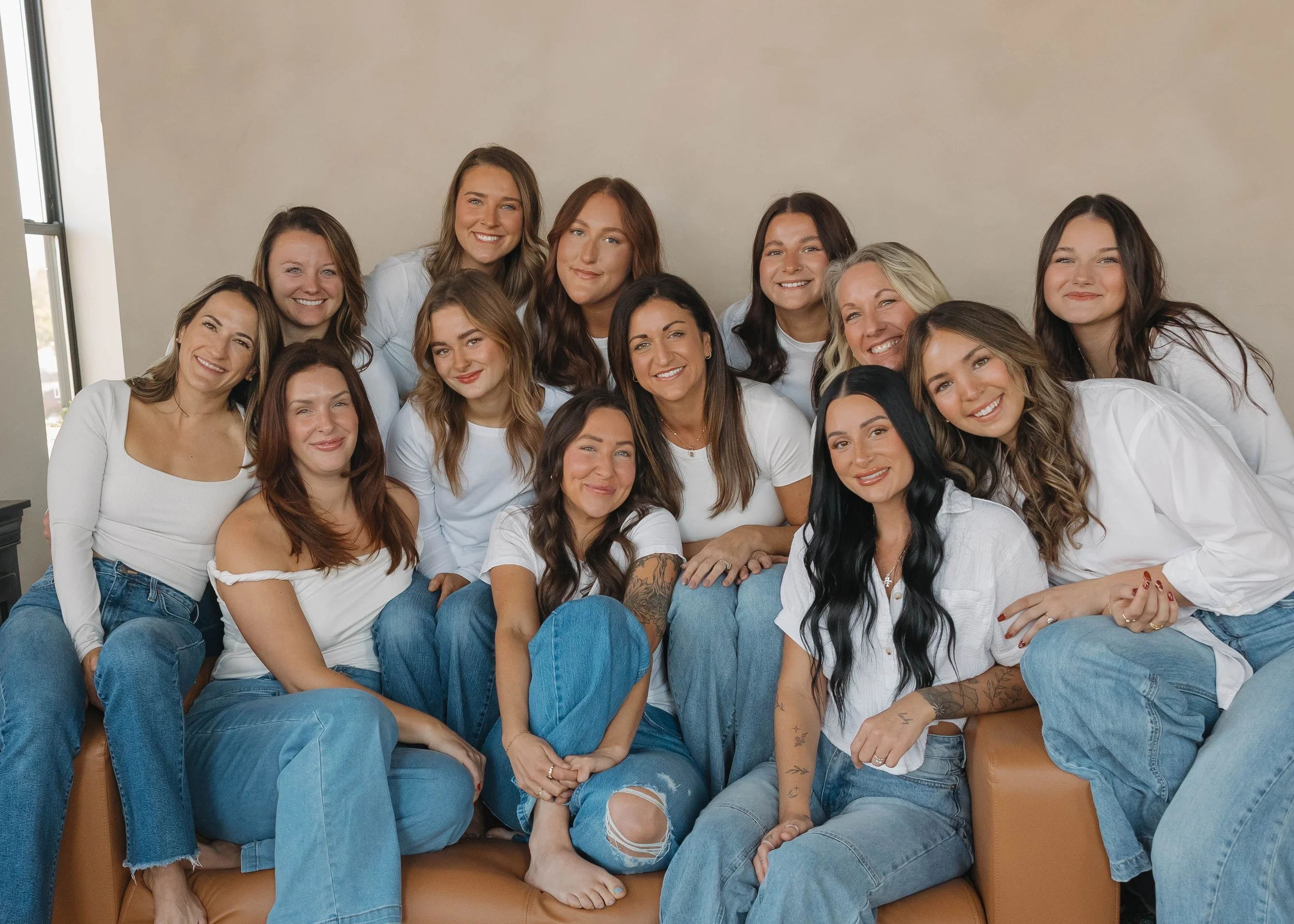 Group of 15 women sitting and standing together indoors, wearing casual white tops and blue jeans, smiling for the camera.