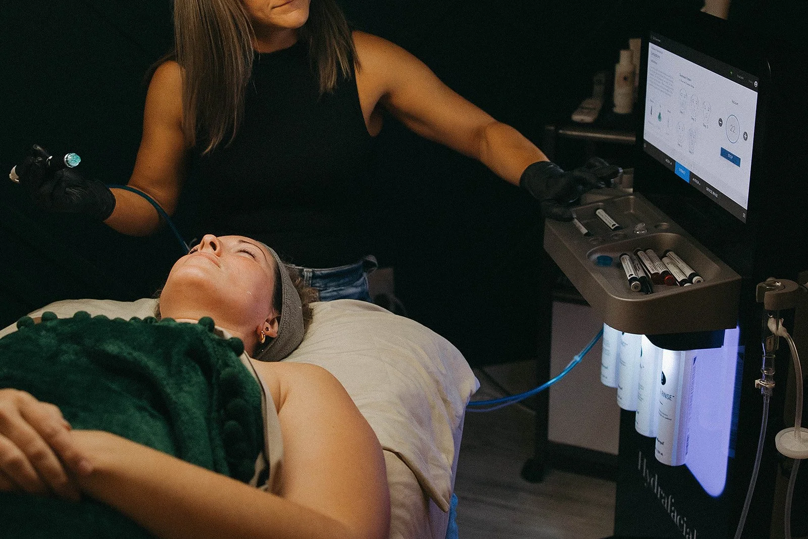 A woman lying on a bed receiving a cosmetic or dermatological treatment from a professional using a laser device with a digital screen in a dark room.