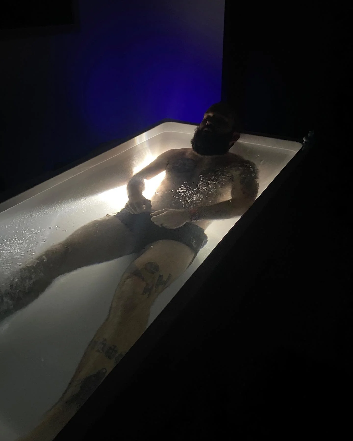A man with a beard and tattoos, wearing black shorts, lying in a lit acrylic tub filled with water in a dark room.