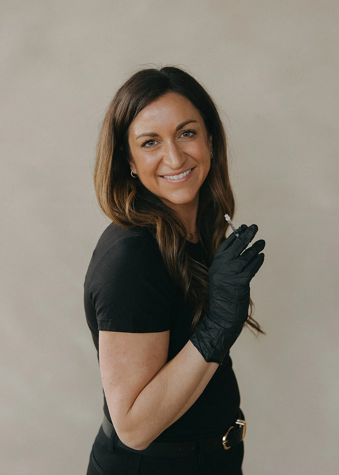 A woman with long brown hair, smiling, dressed in black, wearing black gloves, holding a syringe