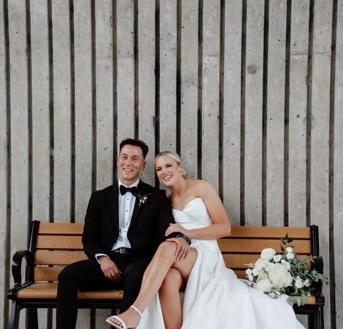 A newlywed couple sitting on a wooden park bench in front of a concrete wall, smiling at the camera. The groom is wearing a black tuxedo with a bow tie, and the bride is in a strapless white wedding dress holding a bouquet of white flowers.