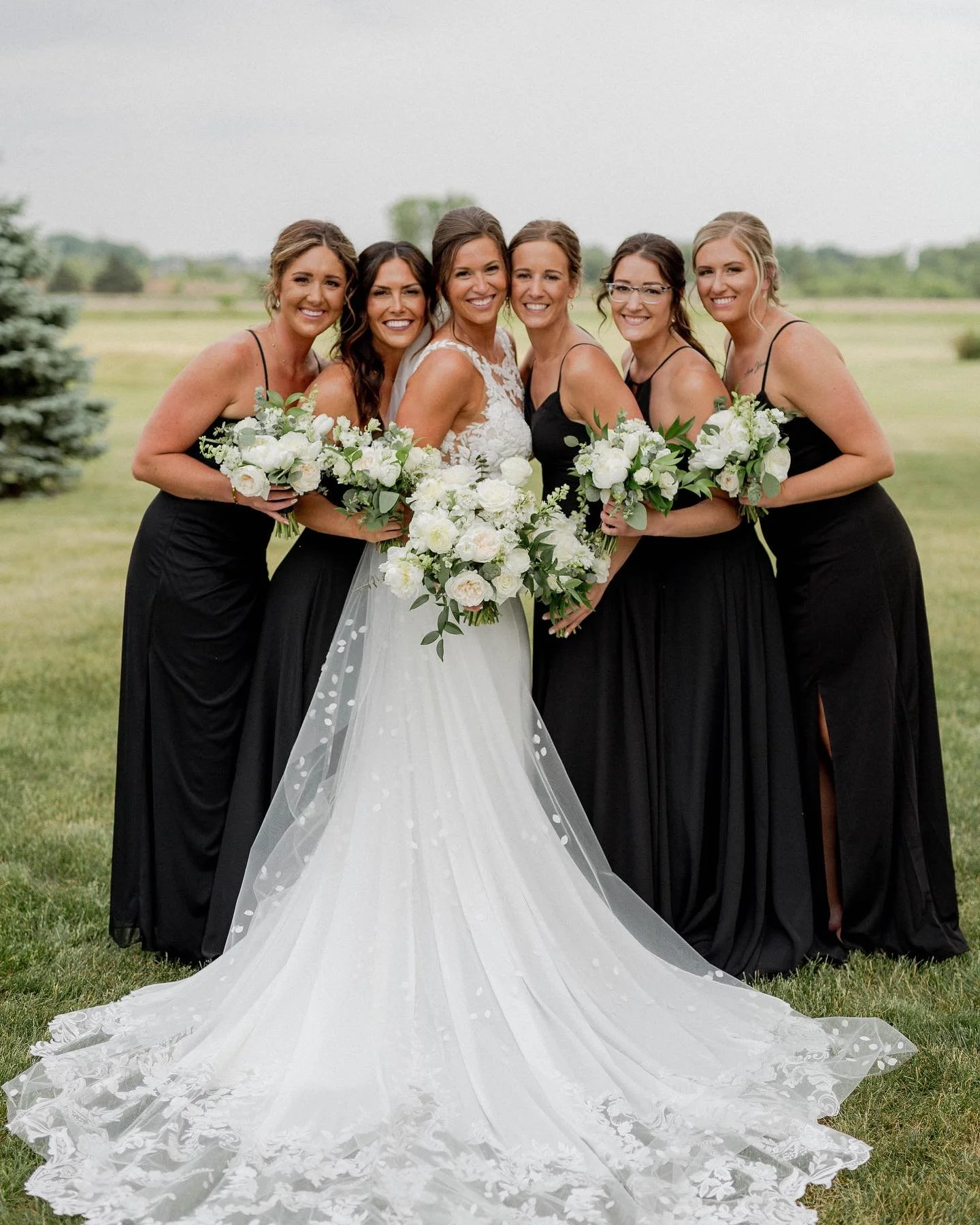 Bride and five bridesmaids in black dresses, holding white bouquets, standing on grass outdoors with a cloudy sky in the background.