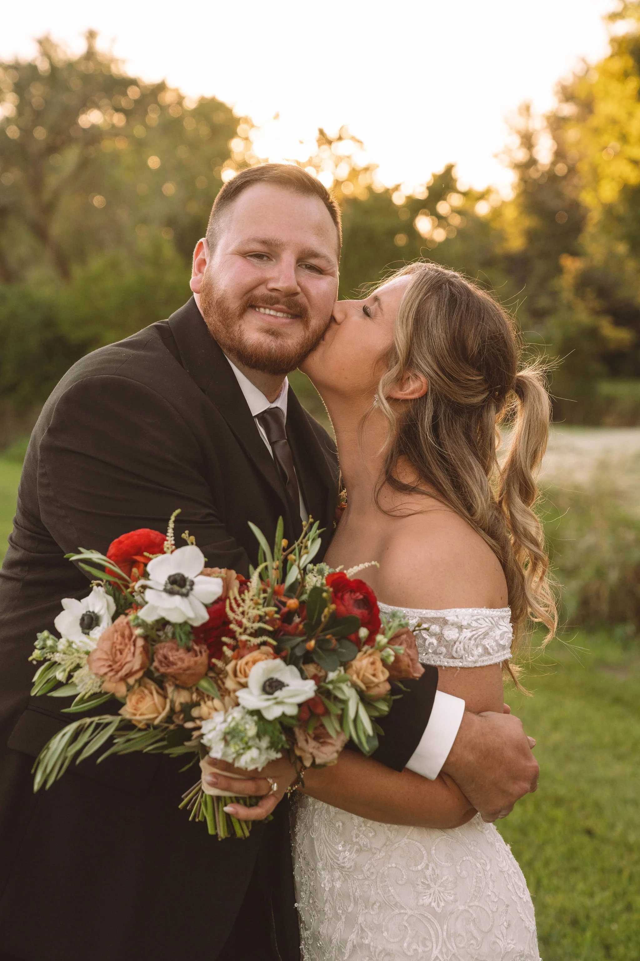 A newlywed couple shares a kiss outdoors during sunset, with the woman holding a bouquet of flowers.