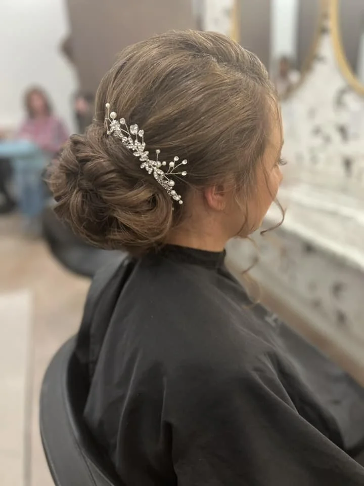 Side view of a woman with an elegant updo hairstyle decorated with a pearl and crystal hair accessory, sitting in a salon.