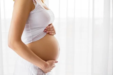 Pregnant woman gently holding her belly in a bright room with sheer curtains.