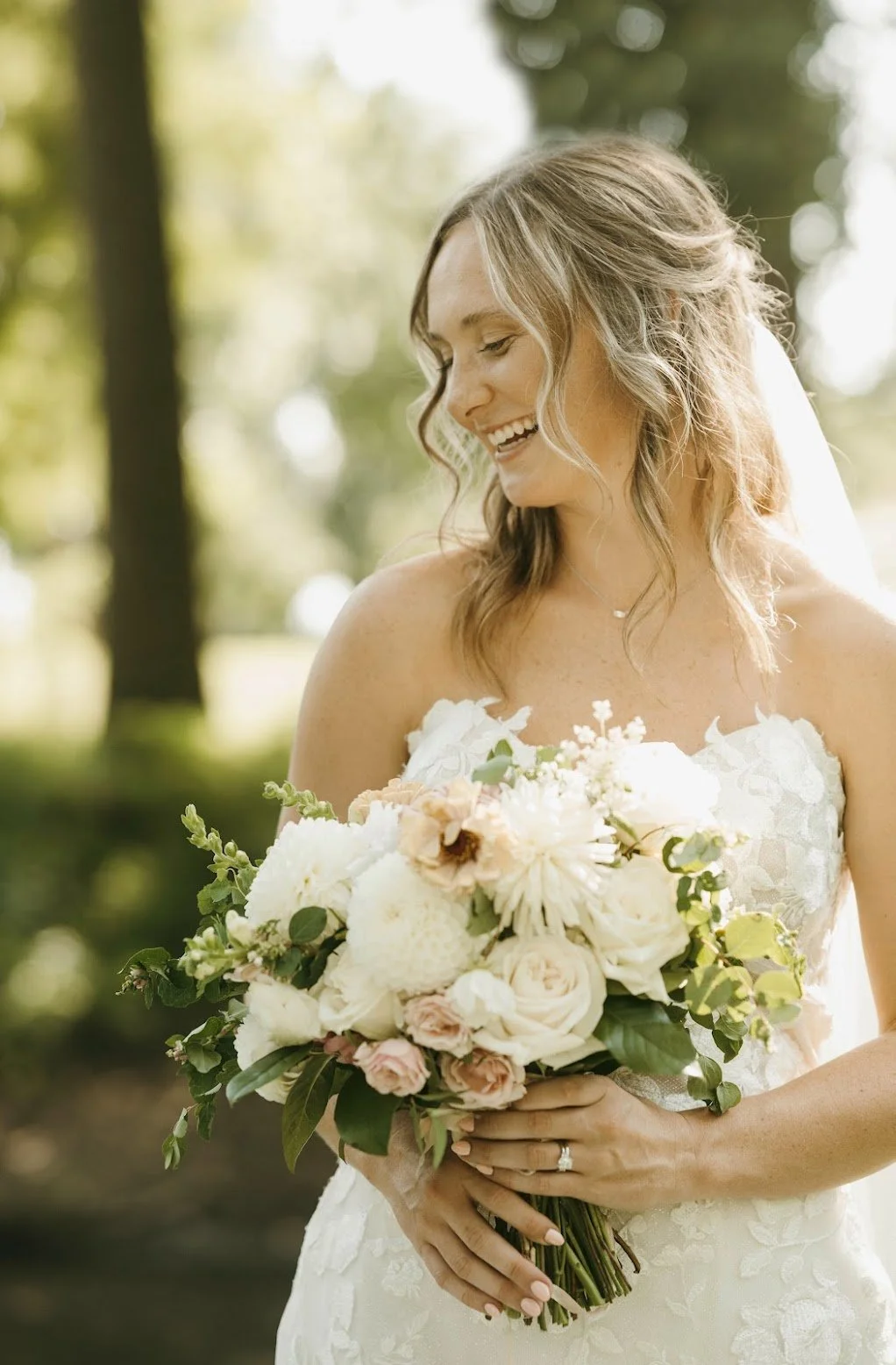 A smiling bride in a white wedding dress holding a bouquet of flowers outdoors, with trees and greenery in the background.