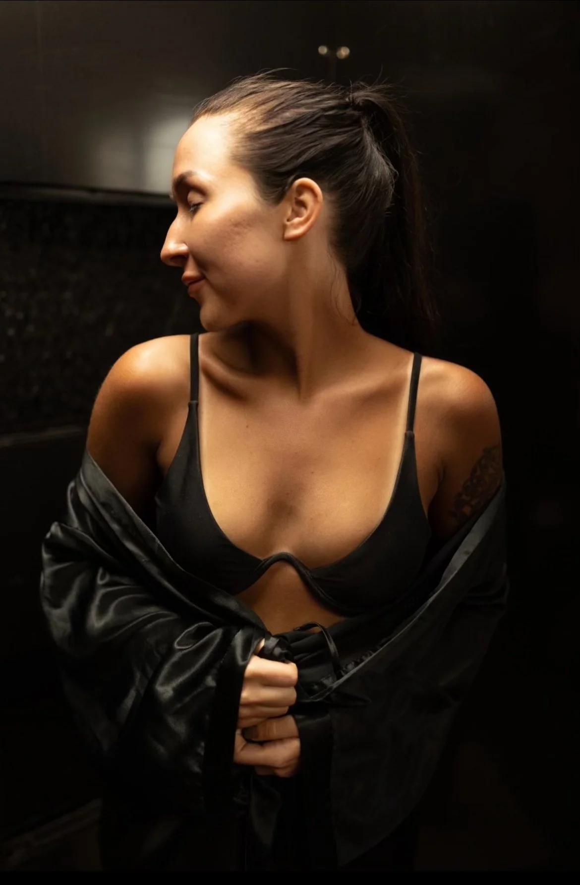 A young woman with dark hair tied back, wearing a black bra and a black jacket, standing against a dark background. She is looking to the side with a slight smile, and her shoulder and collarbone are visible.