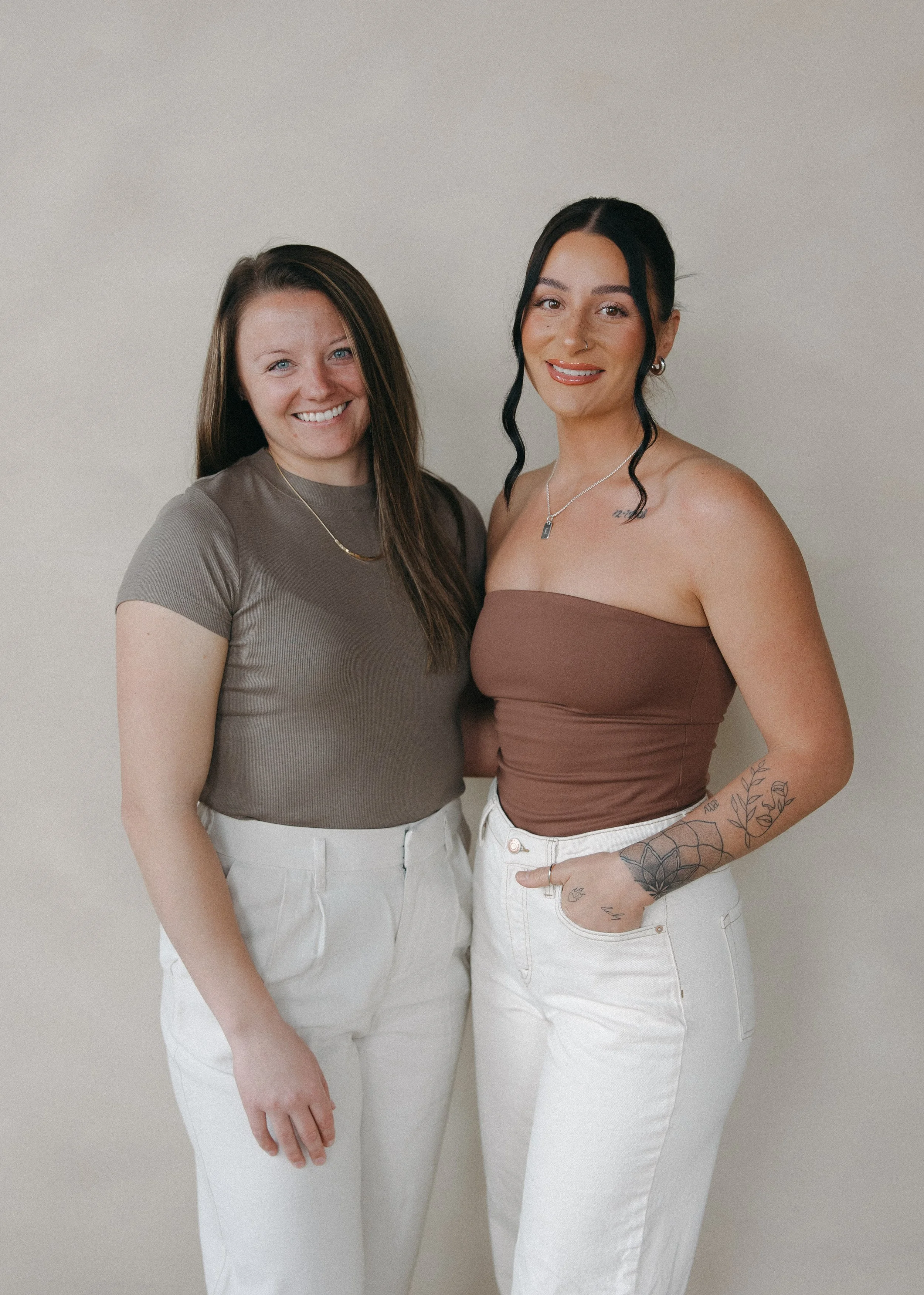Two women standing together against a neutral background, smiling at the camera. One has long brown hair and is wearing a brown t-shirt and white pants. The other has black hair styled in loose waves, visible tattoos on her arm, and is wearing a strapless brown top and white pants.