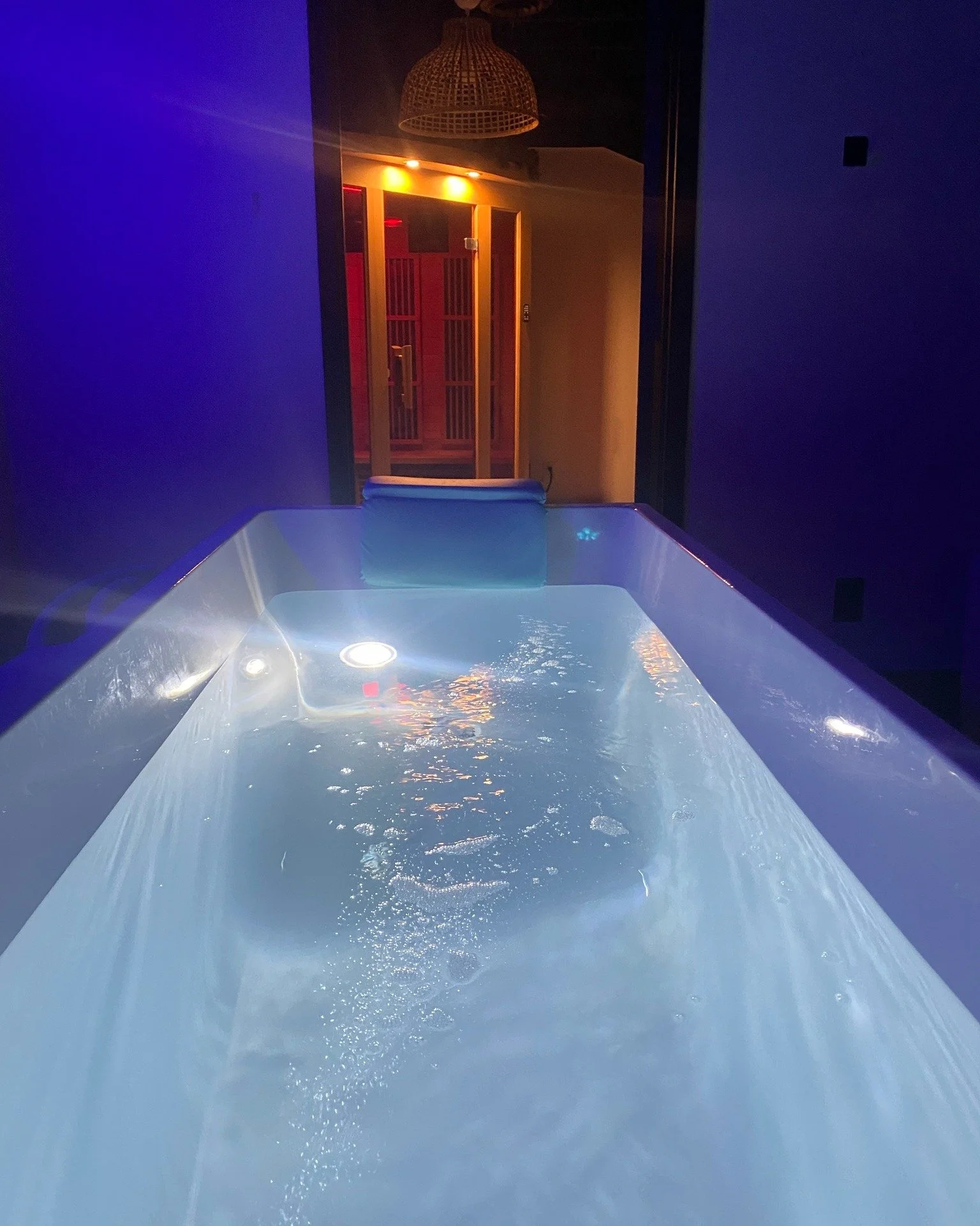 Indoor hot tub filled with water, illuminated by blue LED lights, with a massage pillow attached to the edge, and a door with a red-lit corridor visible in the background.