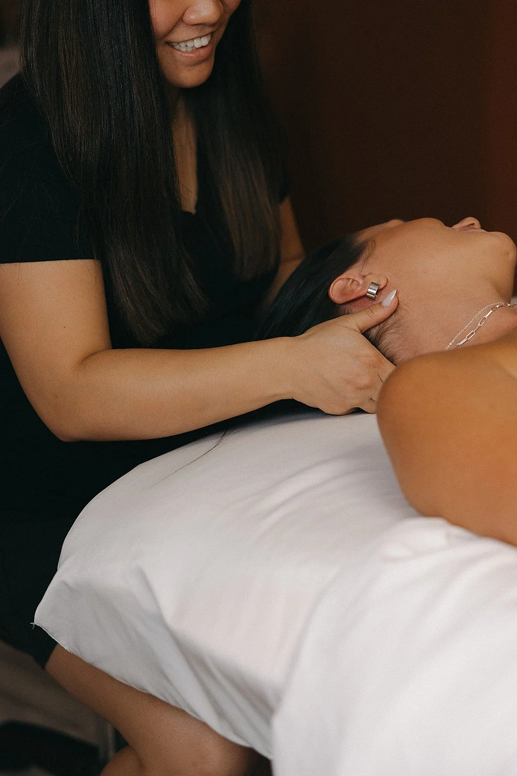 A woman receiving a massage, lying face-up on a massage table, with a massage therapist applying massage techniques to her neck and shoulders.