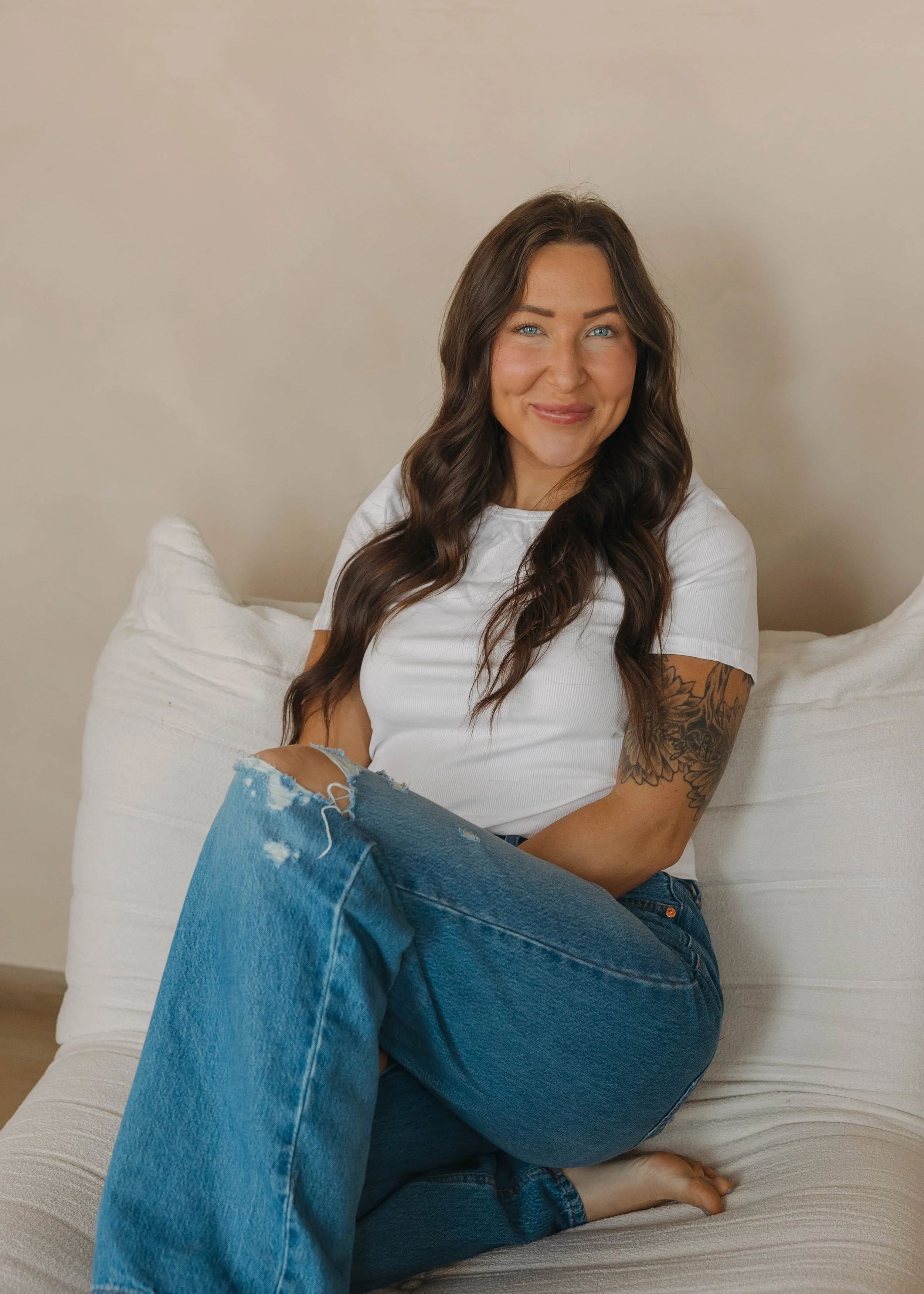 A woman with long wavy brown hair, blue eyes, wearing a white t-shirt and ripped blue jeans, sitting on a white sofa with a beige wall in the background.