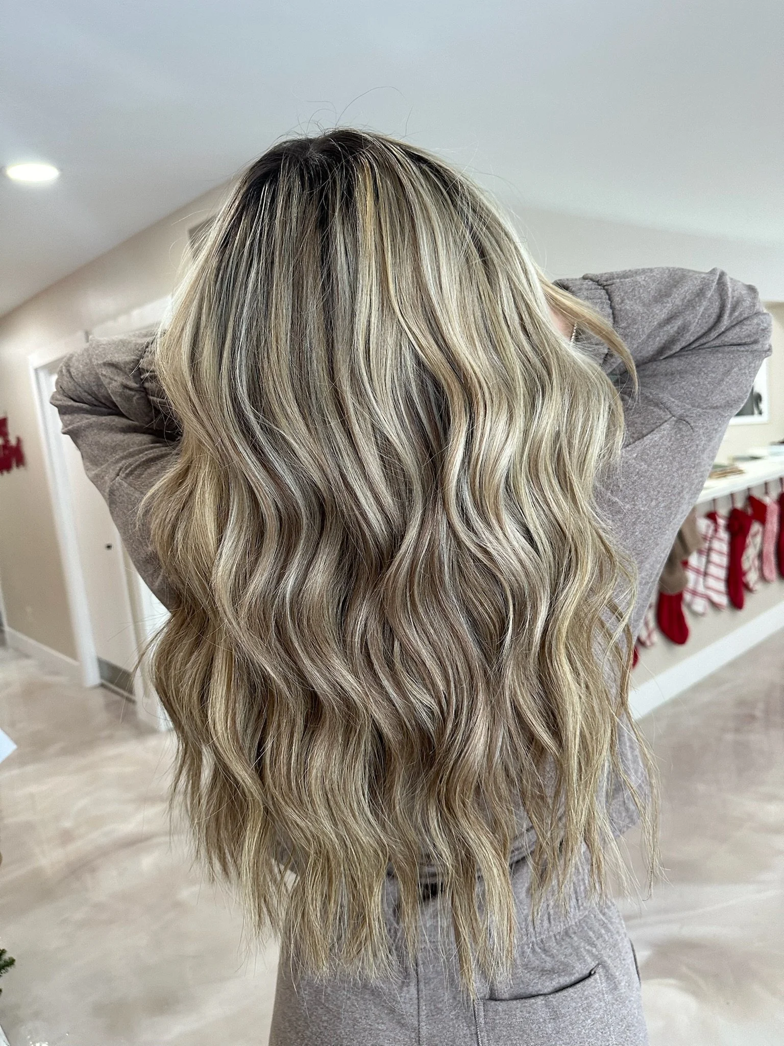 Back of a woman with long, wavy blonde hair, wearing a gray top, in a room decorated for Christmas.