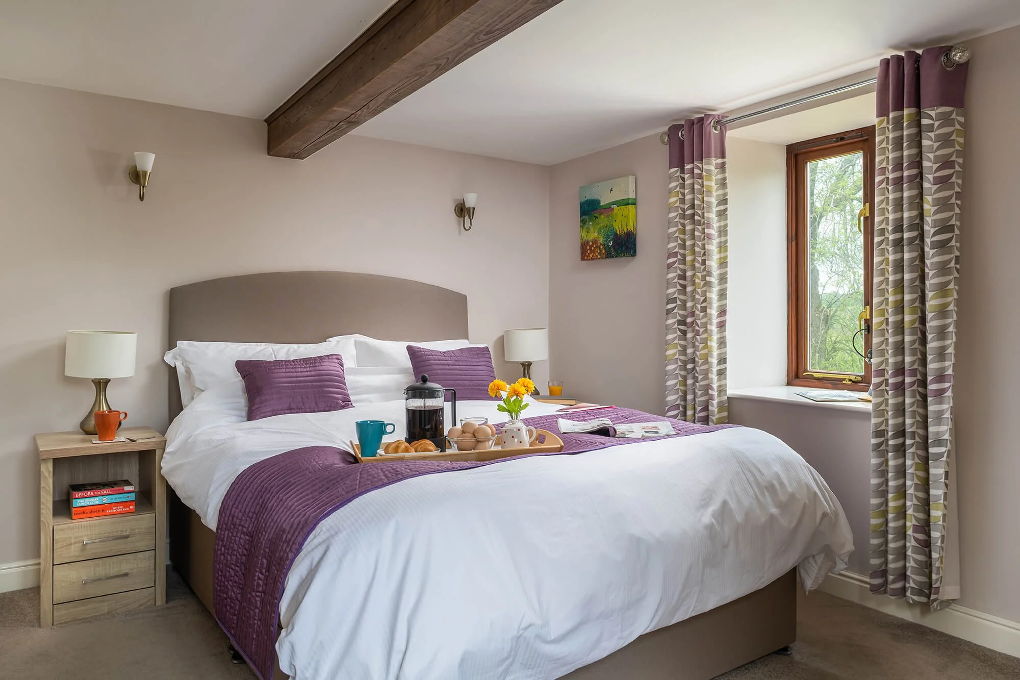 A cosy bedroom with the bed dressed in white sheets and purple pillowcases and purple quilt. There is a tray on the bed with a French press, eggs, croissants, mugs and a yellow flower. Room has a large window with patterned curtains. 