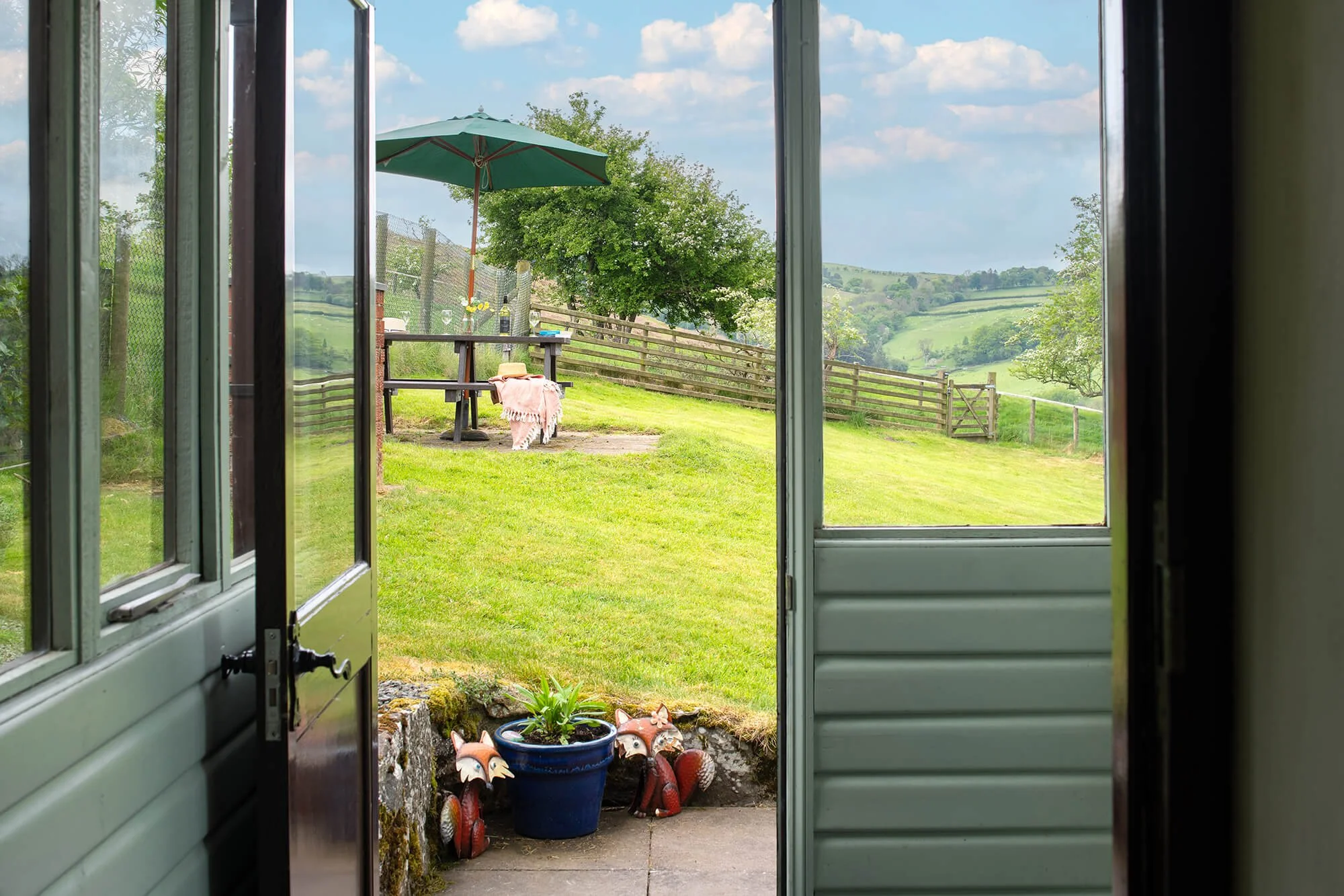 View through an open porch door looking out onto a garden with a bench set for lunch with a green parasol. Trees and hills in the background