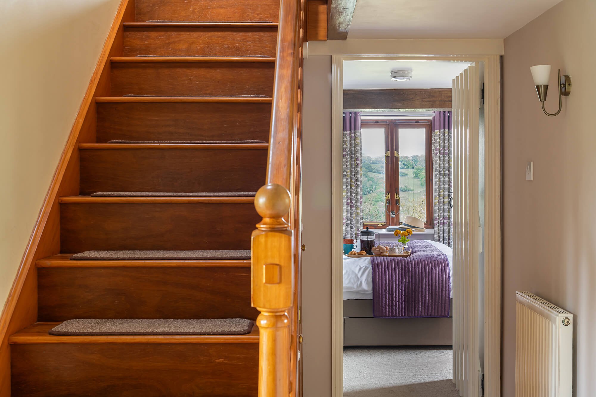 Wooden staircase next to a doorway opening into a bedroom with a purple bedspread, window, and outdoor view.