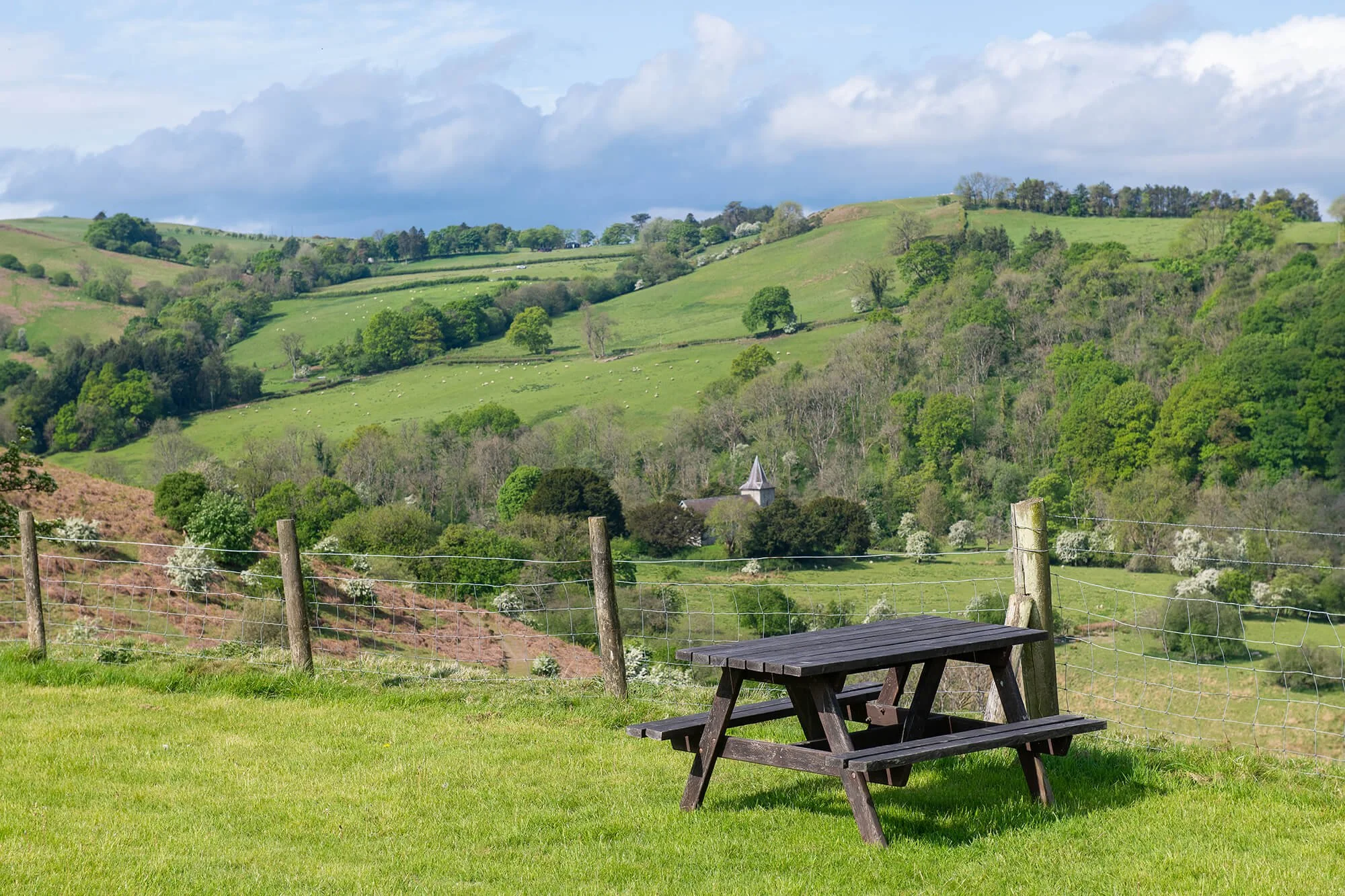 A wooden picnic table on a green grassy hill with a rural landscape of rolling green hills, trees, a church steeple, a wire fence, and a partly cloudy sky in the background.