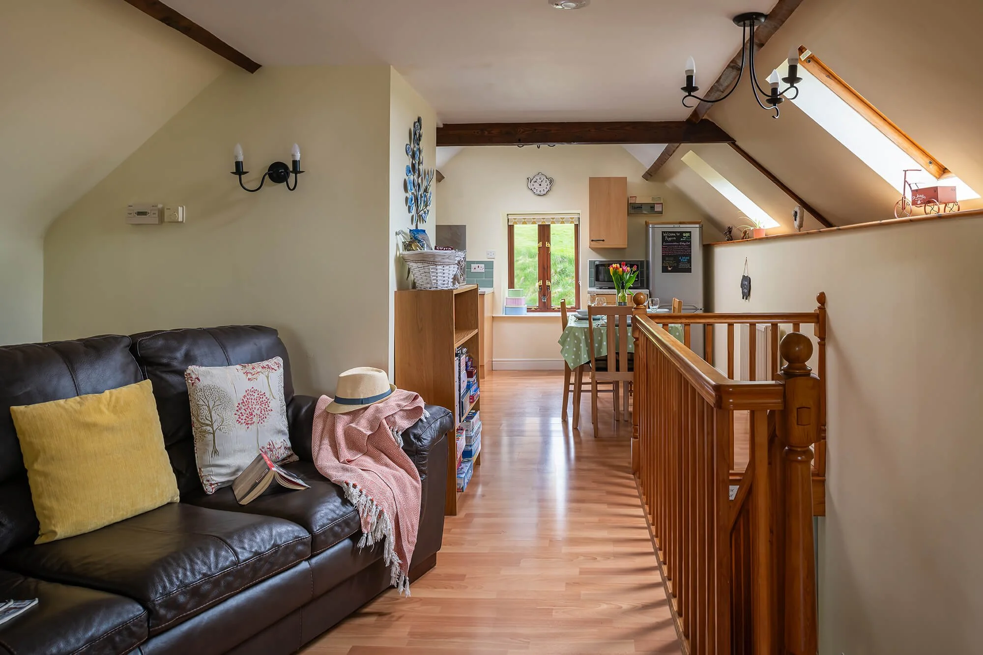 Living area with dark brown sofa, cushions, blanket with a hat on top. Wooden staircase with railings on the right side, skylight illuminates the area. In the background there is a dining table and chairs
