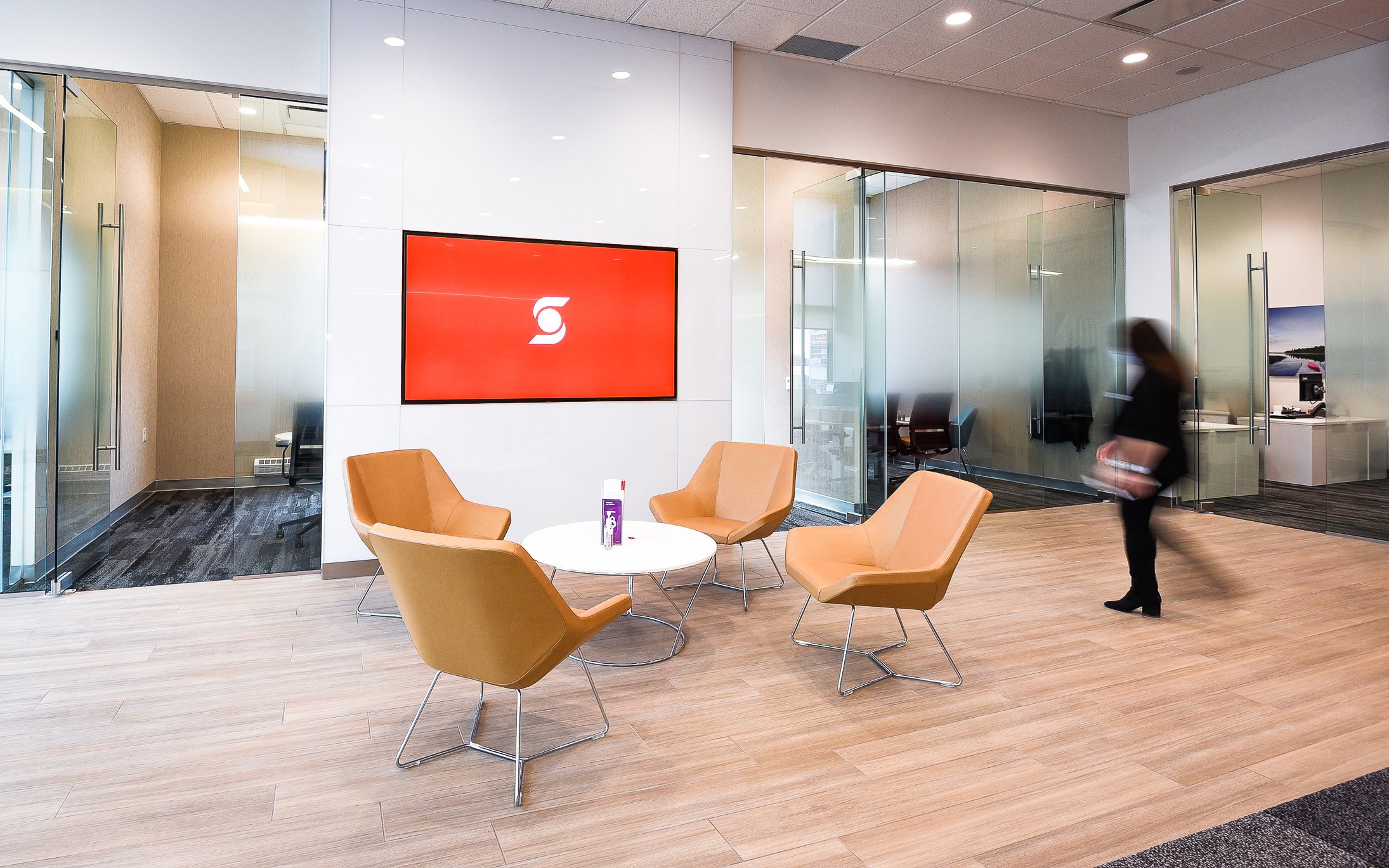 Modern office lobby with four orange chairs around a white round table, a large digital screen on the wall displaying a red background with a white logo, glass walls and doors leading to conference rooms, and a woman walking by holding a tablet.