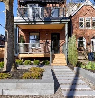 Front view of a brick house with a small porch, stairs leading up to the door, and a front yard with plants and a sidewalk.