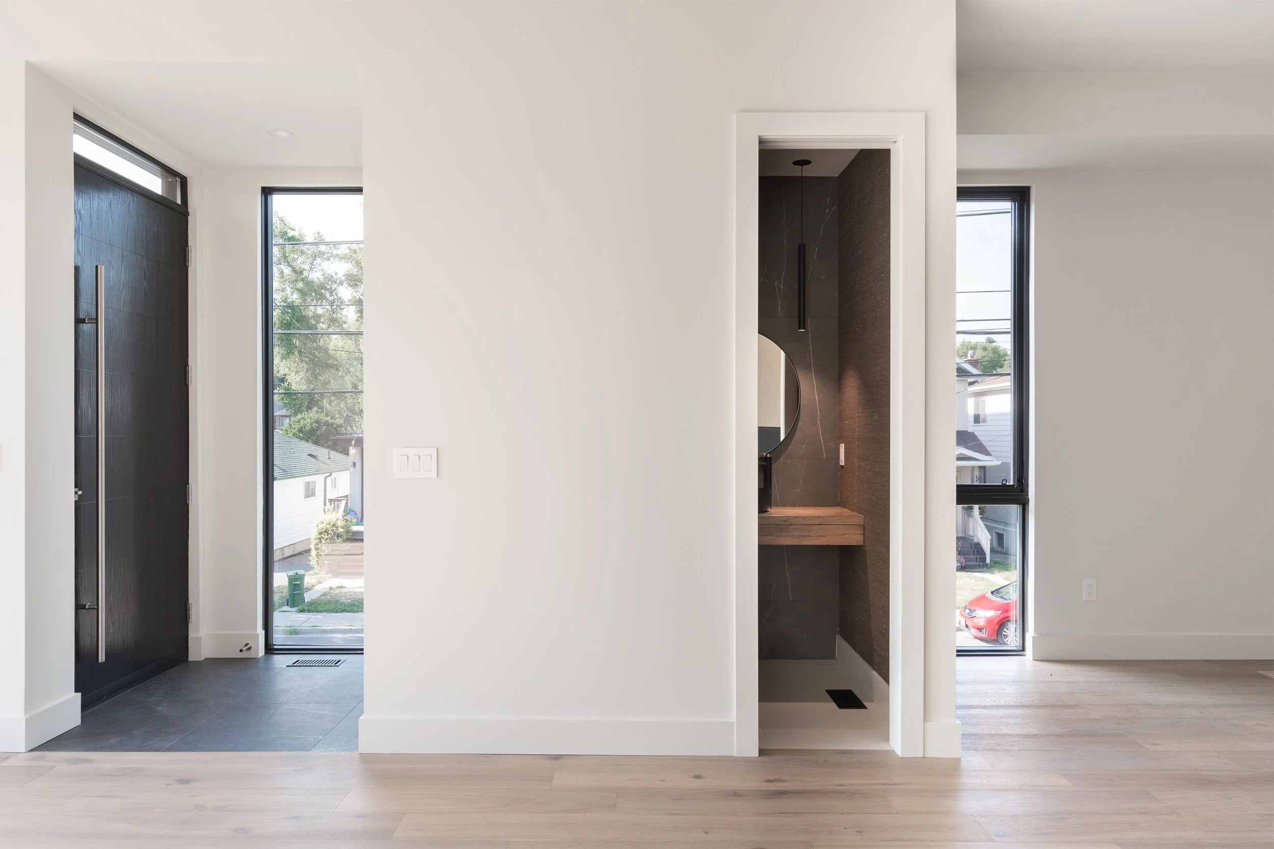 Interior view of a modern home with entrance, hallway, and a small bathroom or closet with dark tiles, round mirror, and wooden countertop.