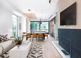 Modern open-concept living and dining room with white walls, large windows, a beige sofa, and a wall-mounted TV.
