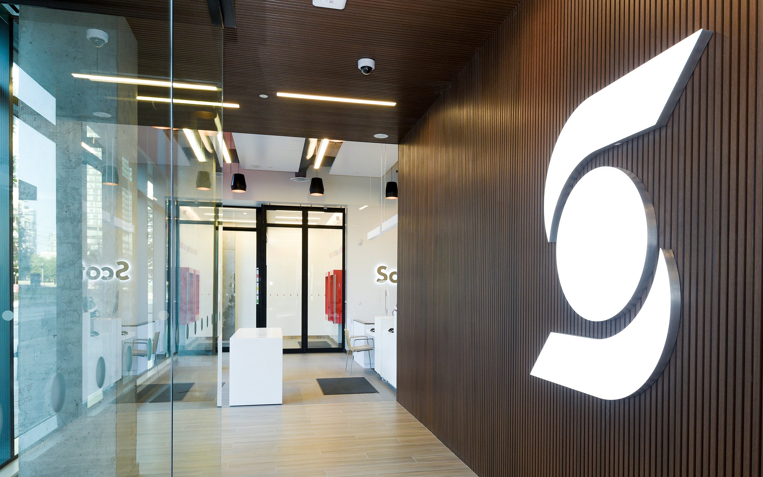 Reception area of an office with a large illuminated logo on a wooden wall, glass walls with reflections, and modern lighting fixtures.