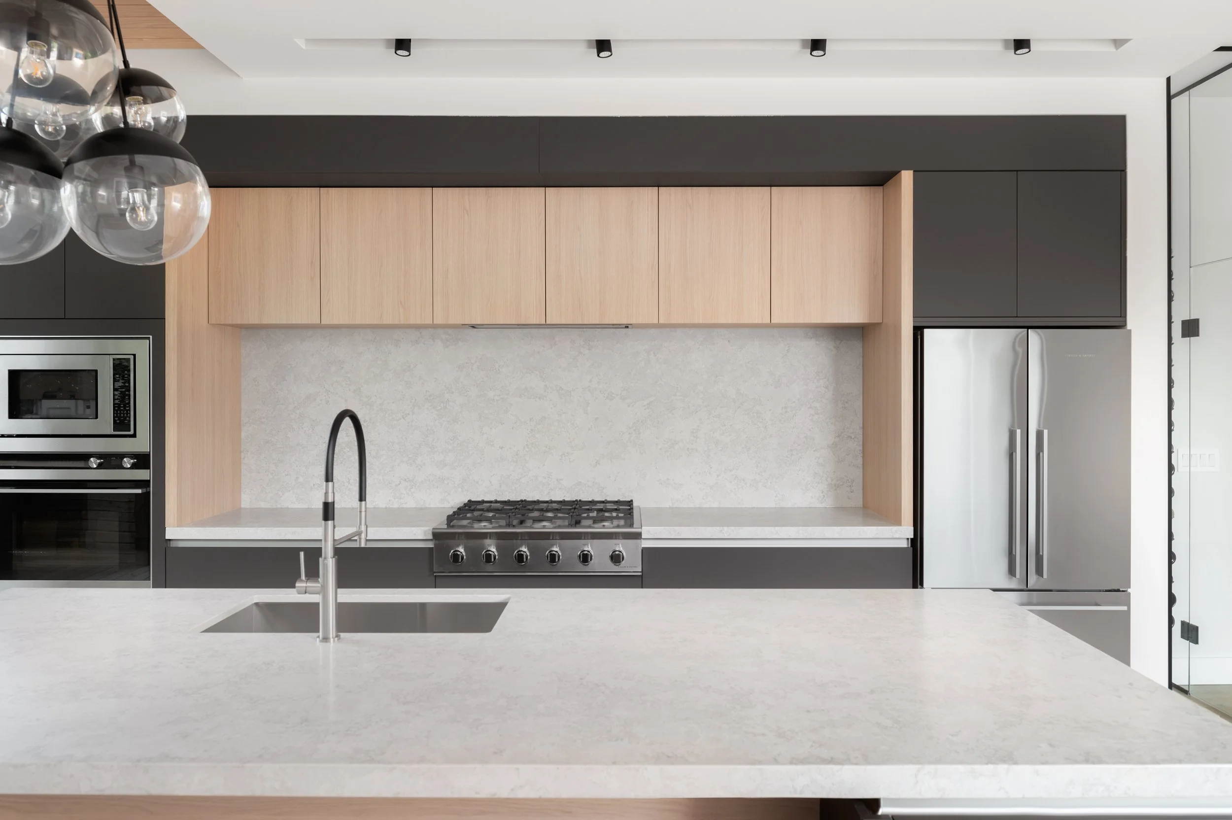 Modern kitchen with light wood and black cabinetry, stainless steel refrigerator, built-in oven and microwave, black and stainless steel gas stove, and a large white countertop island with a sink.