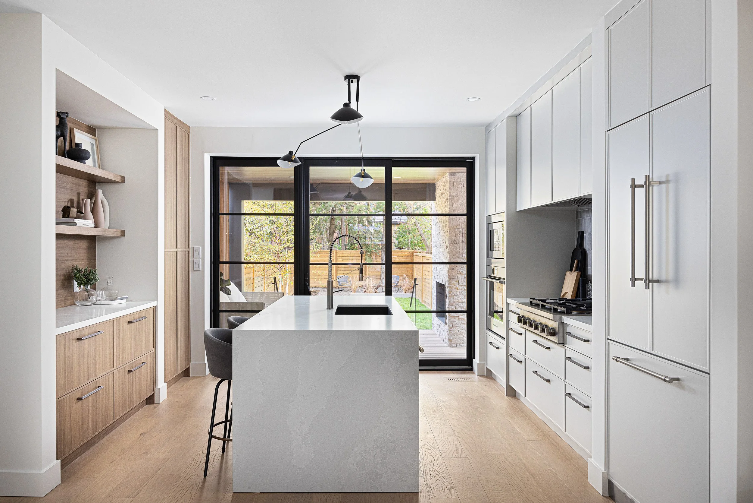 Modern kitchen with white cabinetry, a central island with a sink, black framed glass sliding doors, wooden flooring, and a view of a backyard with a patio and trees.