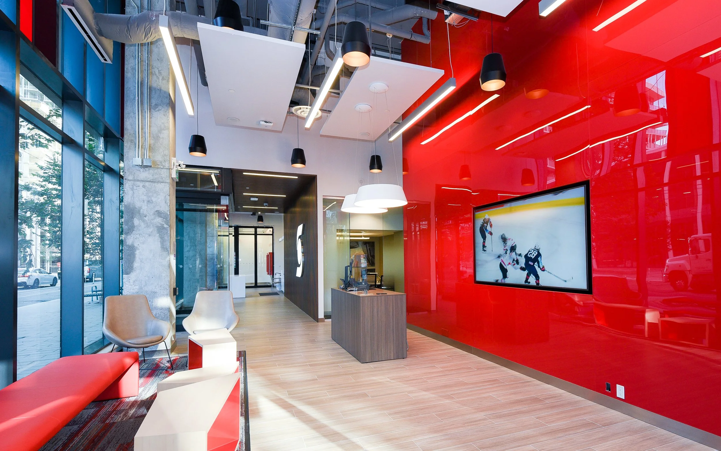 Modern office reception area with red accent wall, large screen displaying hockey game, glass front, and stylish chairs.