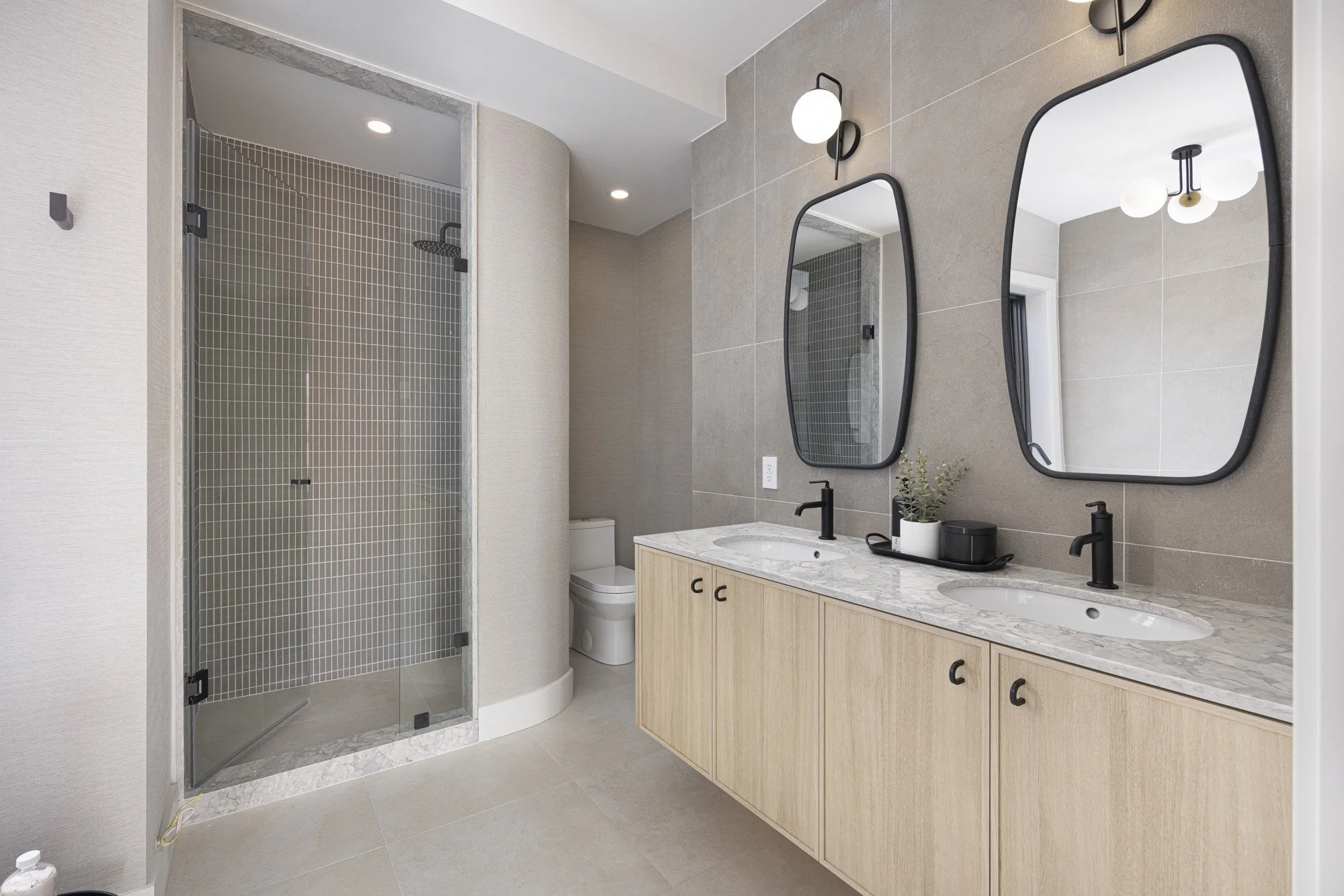 Modern bathroom with a double vanity with oval mirrors, black fixtures, and a marble countertop, a tiled shower with a glass door, and a toilet in the background.