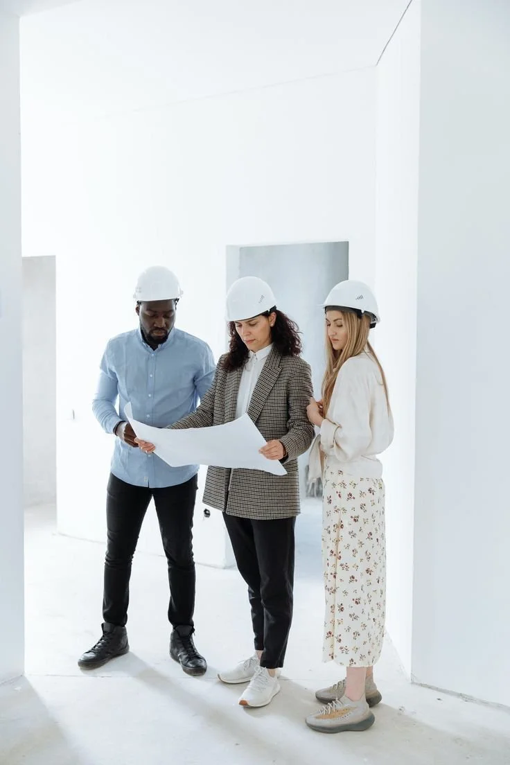 Three construction workers wearing white safety helmets looking at blueprints inside an unfinished white room.