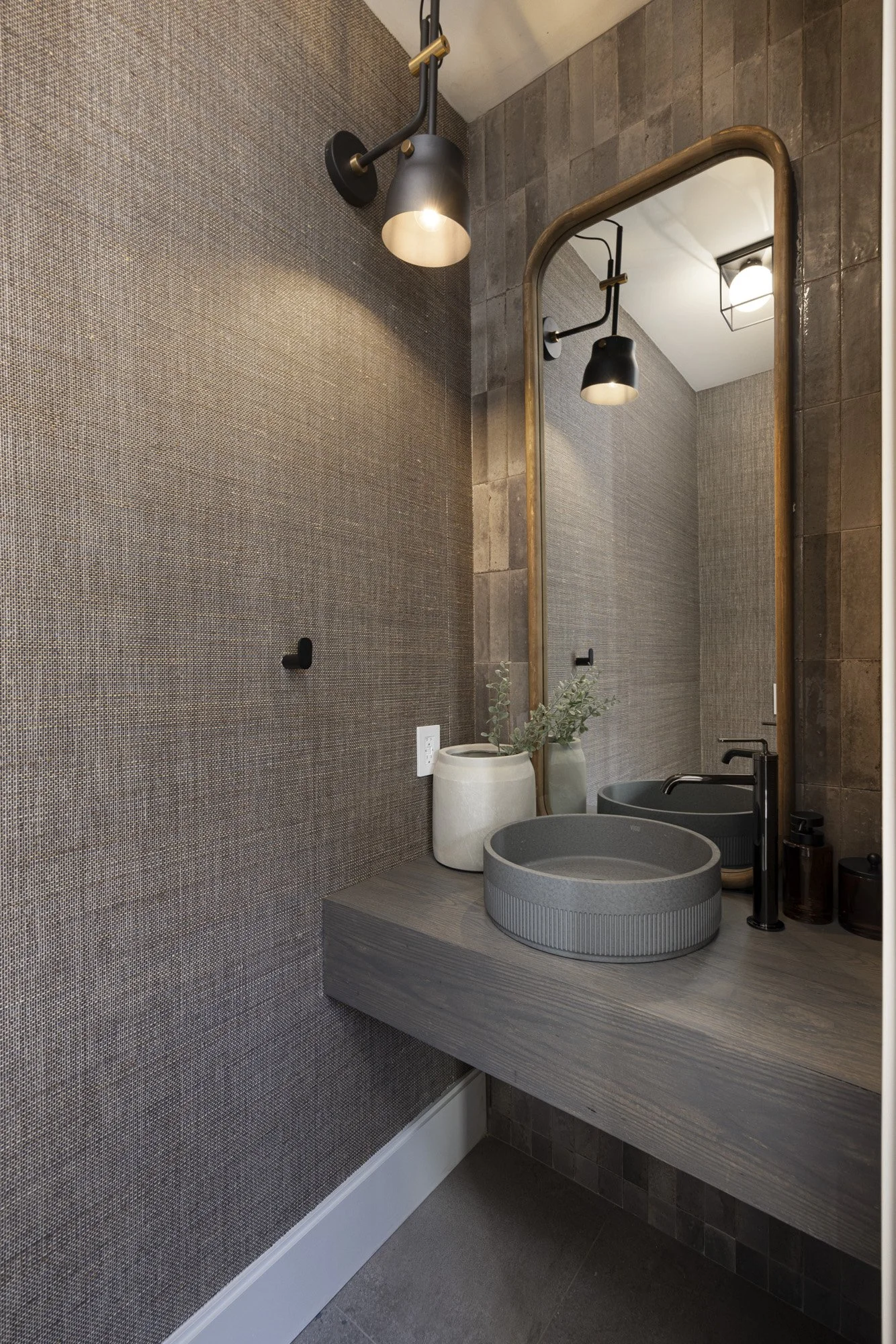 Modern bathroom with textured brown wall, a large mirror, black and brass light fixtures, a gray vanity with a round vessel sink, and minimalist decor including vases and soap dispensers.