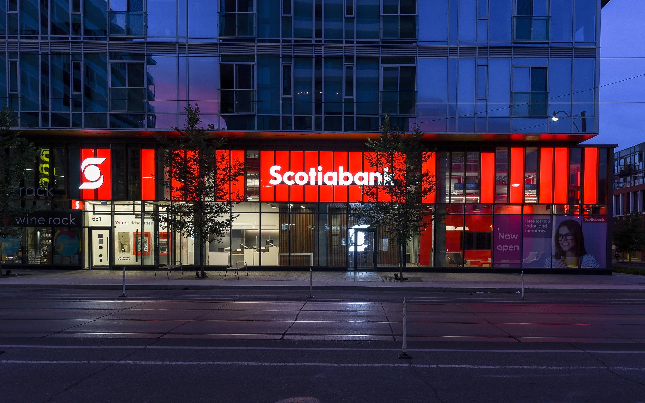 Night view of a Scotiabank branch with a bright red illuminated sign and window accents, situated on a city street with tram tracks in the foreground and trees outside.