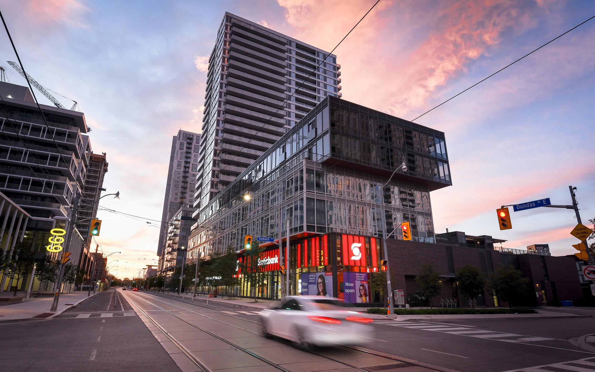 City street scene at dusk with modern buildings, traffic lights, and a blurred white car moving through.