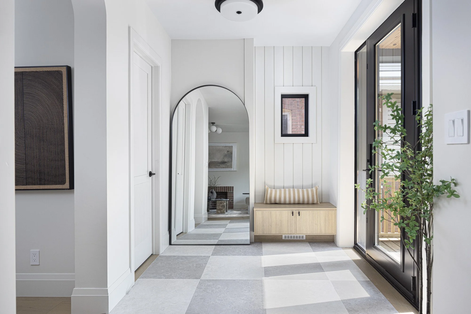 Modern entrance or hallway with a door opening to an outside deck, a small bench with a striped pillow, a tall plant, a large mirror, and minimalist decor.