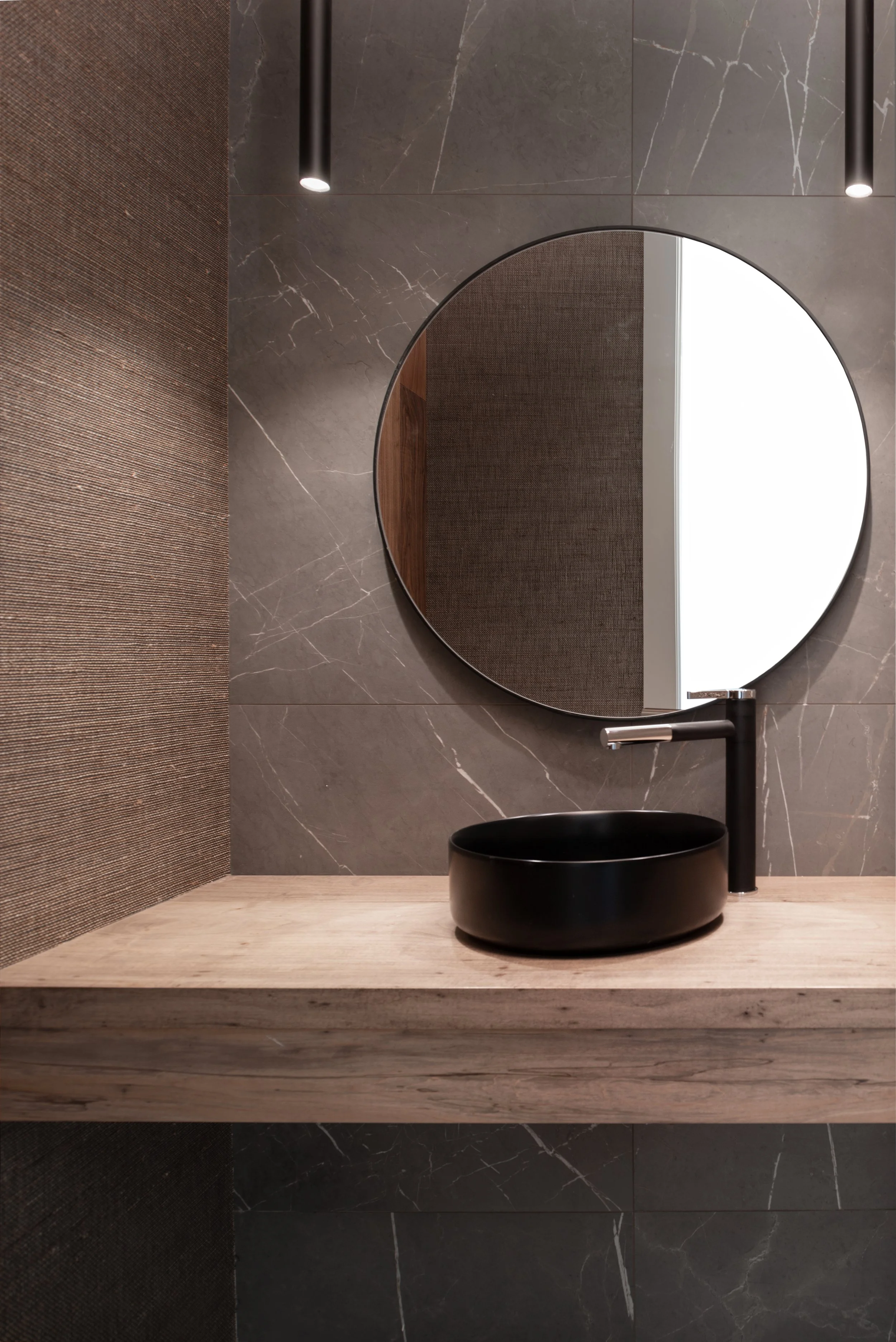 Modern bathroom sink with a black vessel basin, black faucet, round mirror, gray marble wall, textured brown wall, and light wooden countertop.