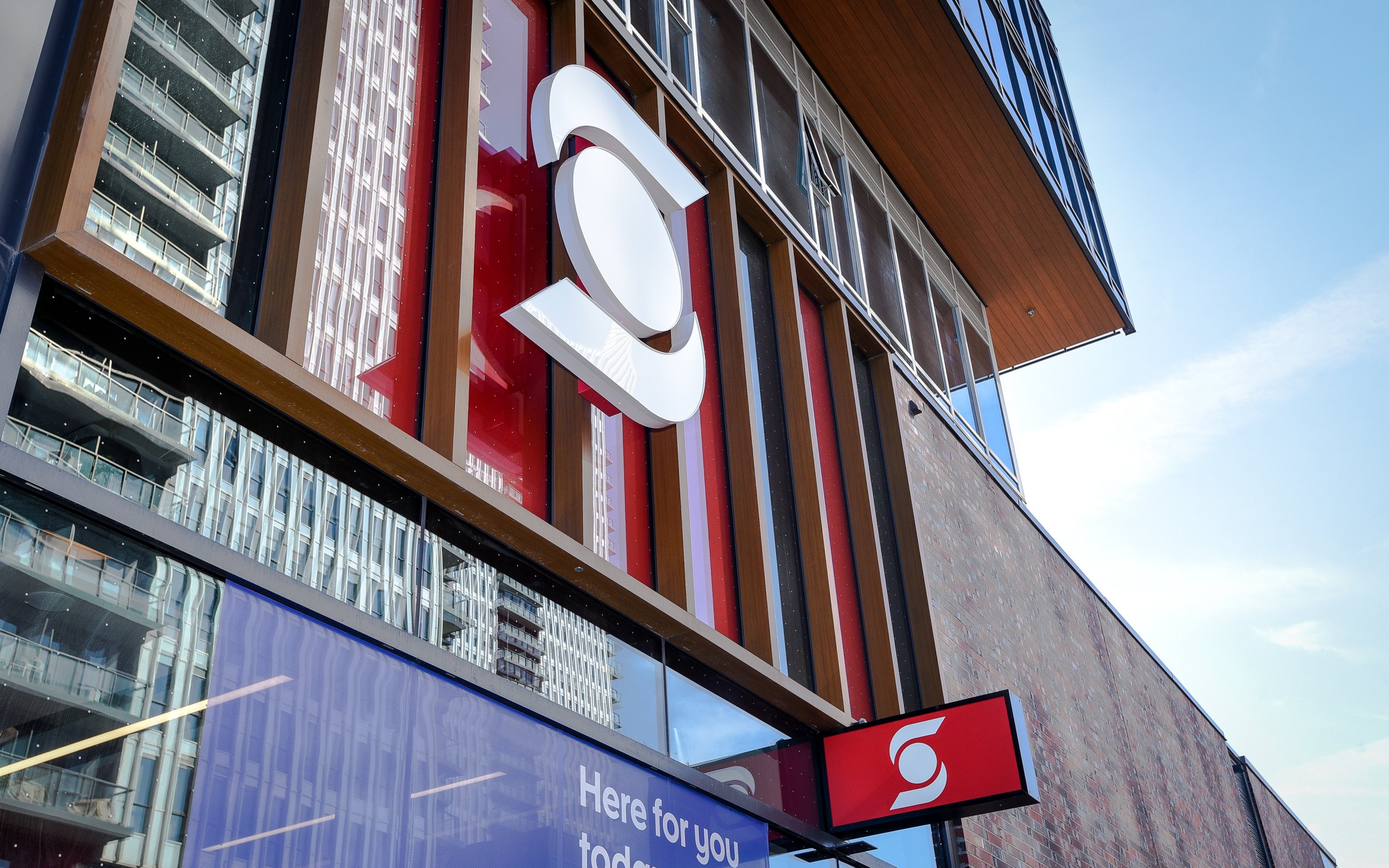 The exterior of a modern shopping mall or retail store with large red and white signage and a digital billboard displaying a message.